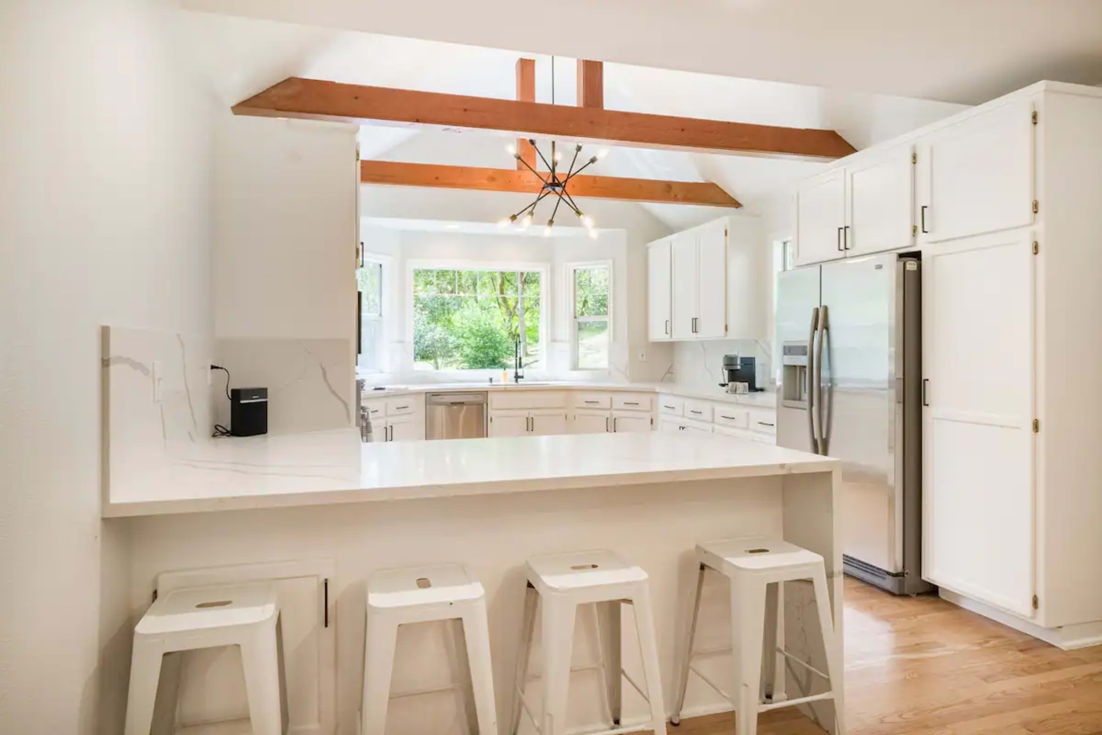 A modern kitchen with white cabinets, a central island with bar stools, and wooden beams overhead.