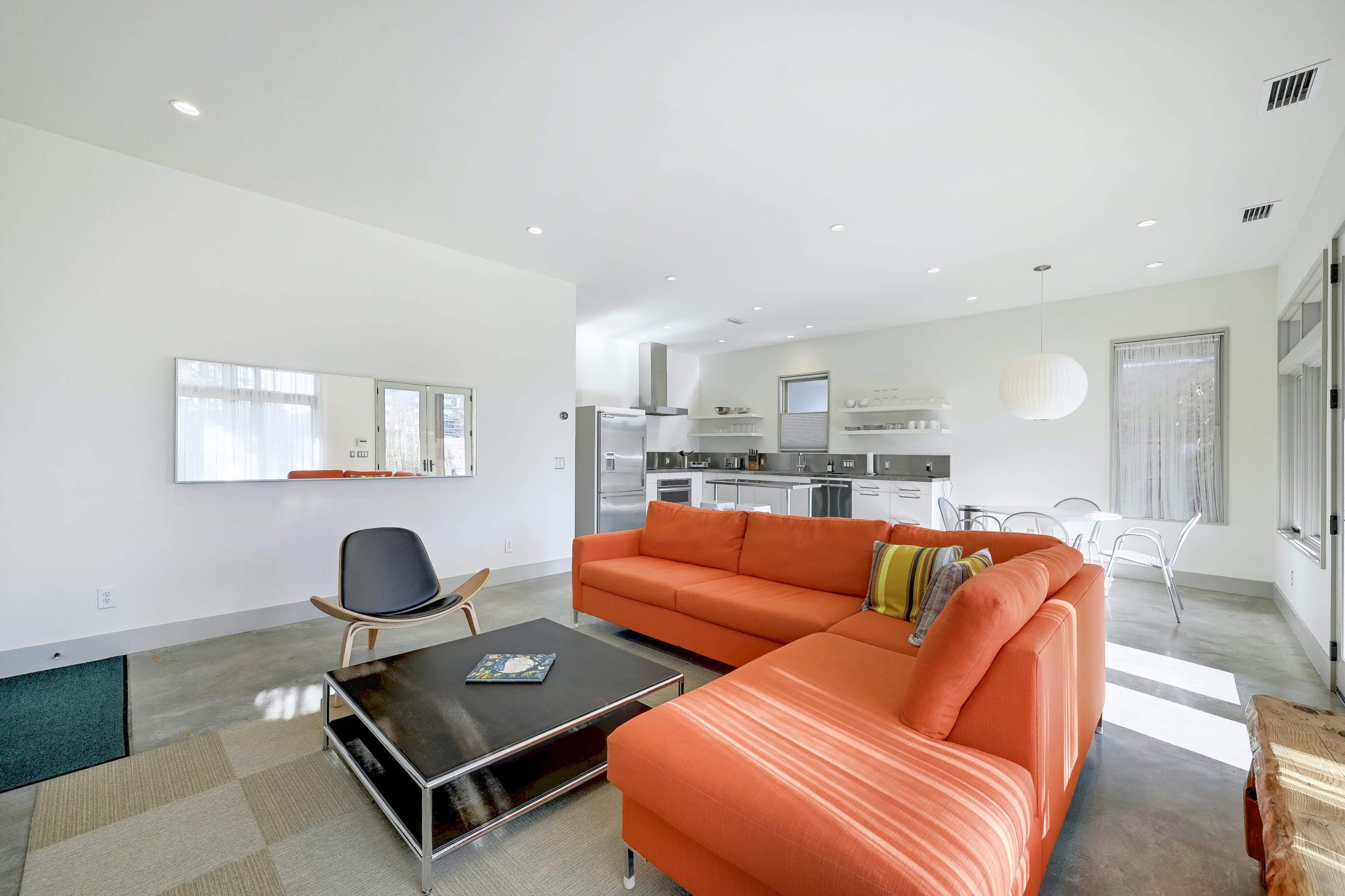 The image shows a modern living space featuring an orange sectional sofa, a glass coffee table, and a kitchen visible in the background.