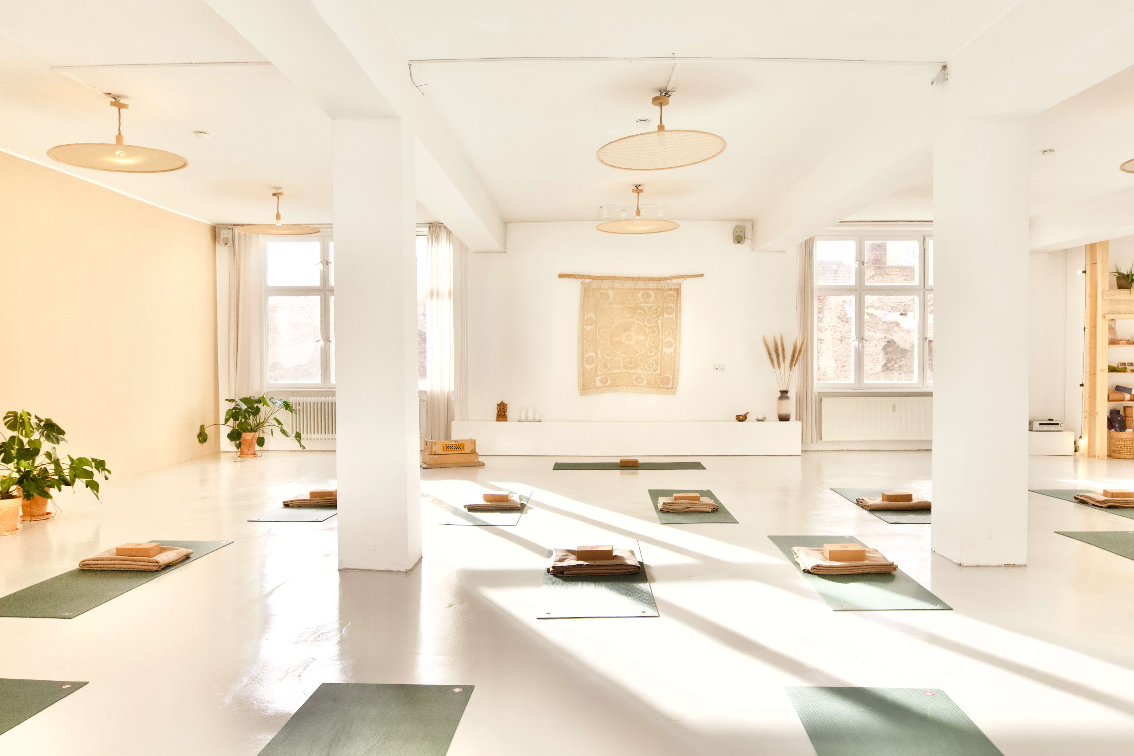 A bright, open yoga studio with rows of mats arranged on a polished white floor, complemented by plants and minimalistic decor.