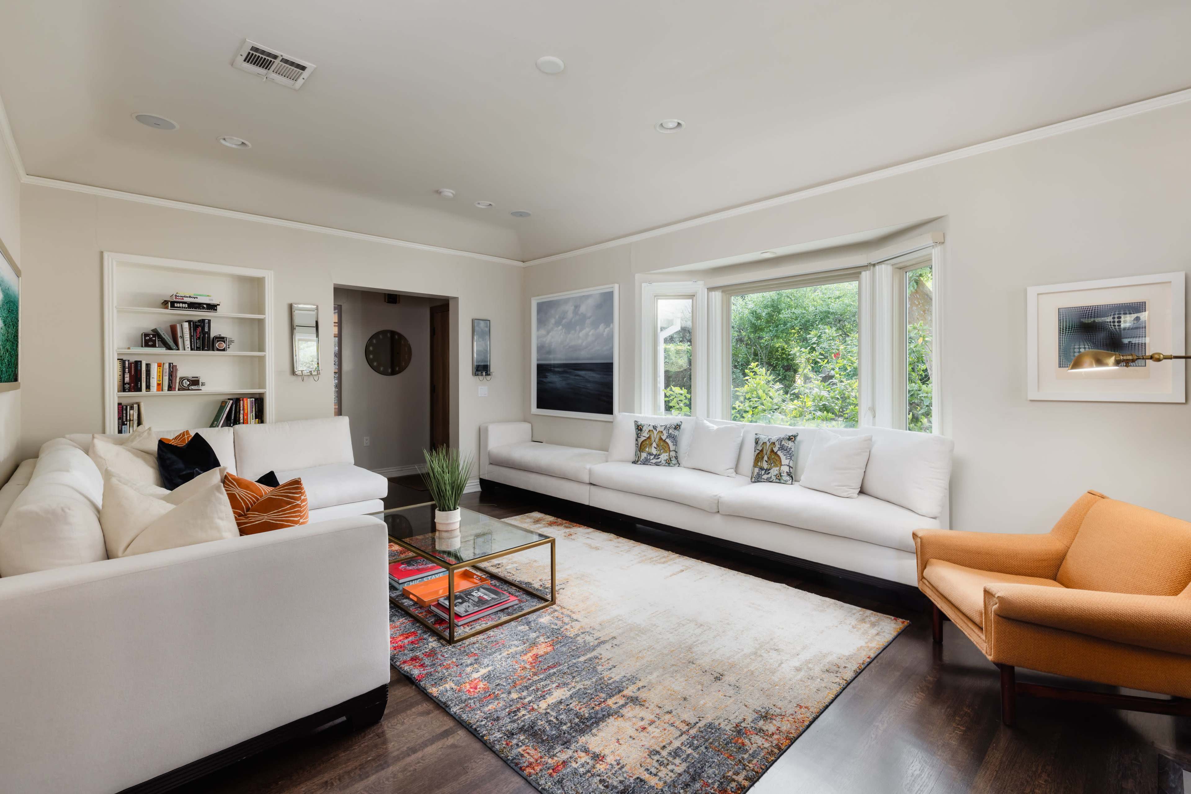 The living room features a white sectional sofa, an orange armchair, a glass coffee table, and a large area rug, with windows offering a view of greenery outside.