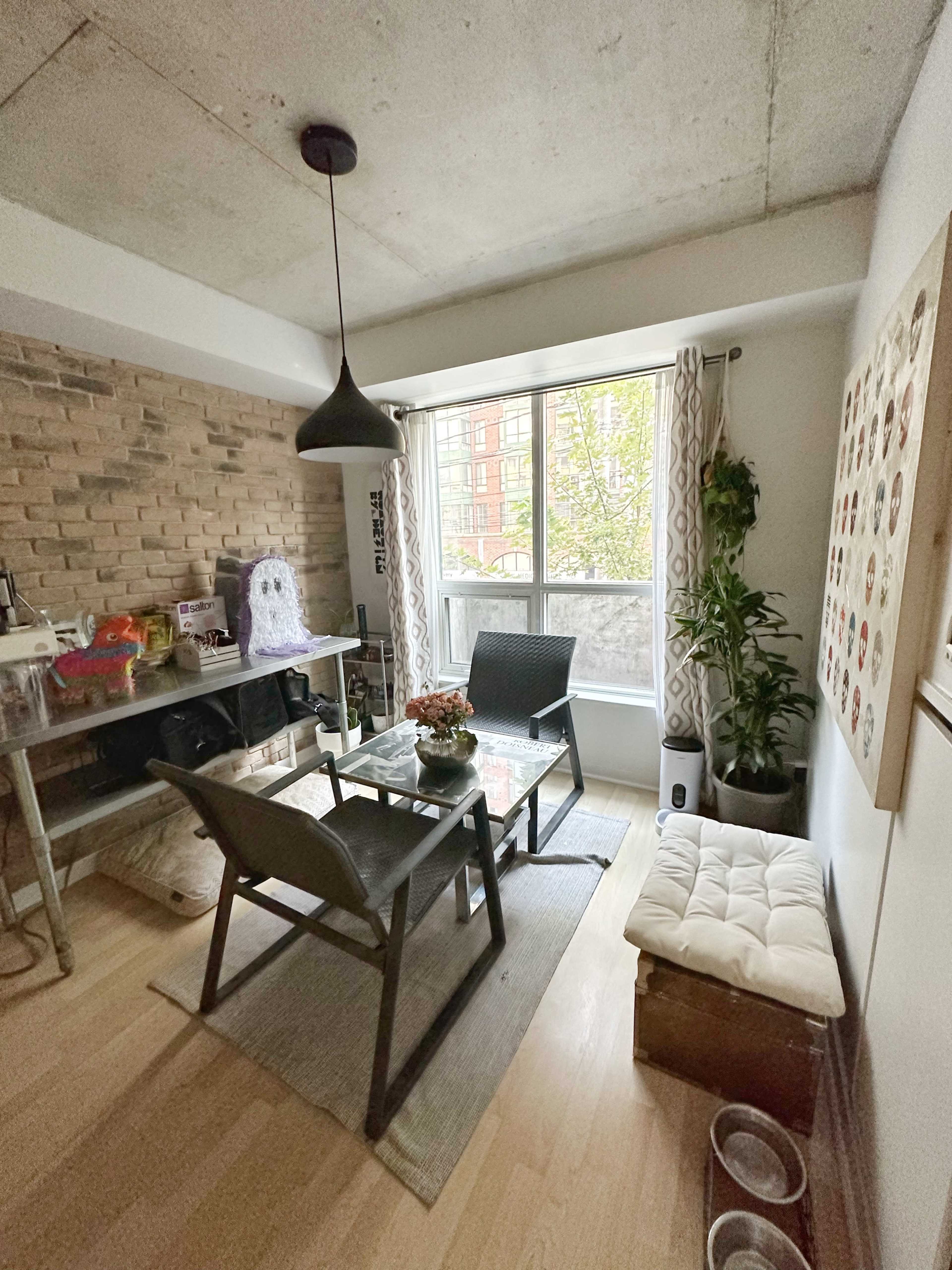 A small dining area features a glass table surrounded by four chairs, with a large window providing natural light and a plant in the corner.