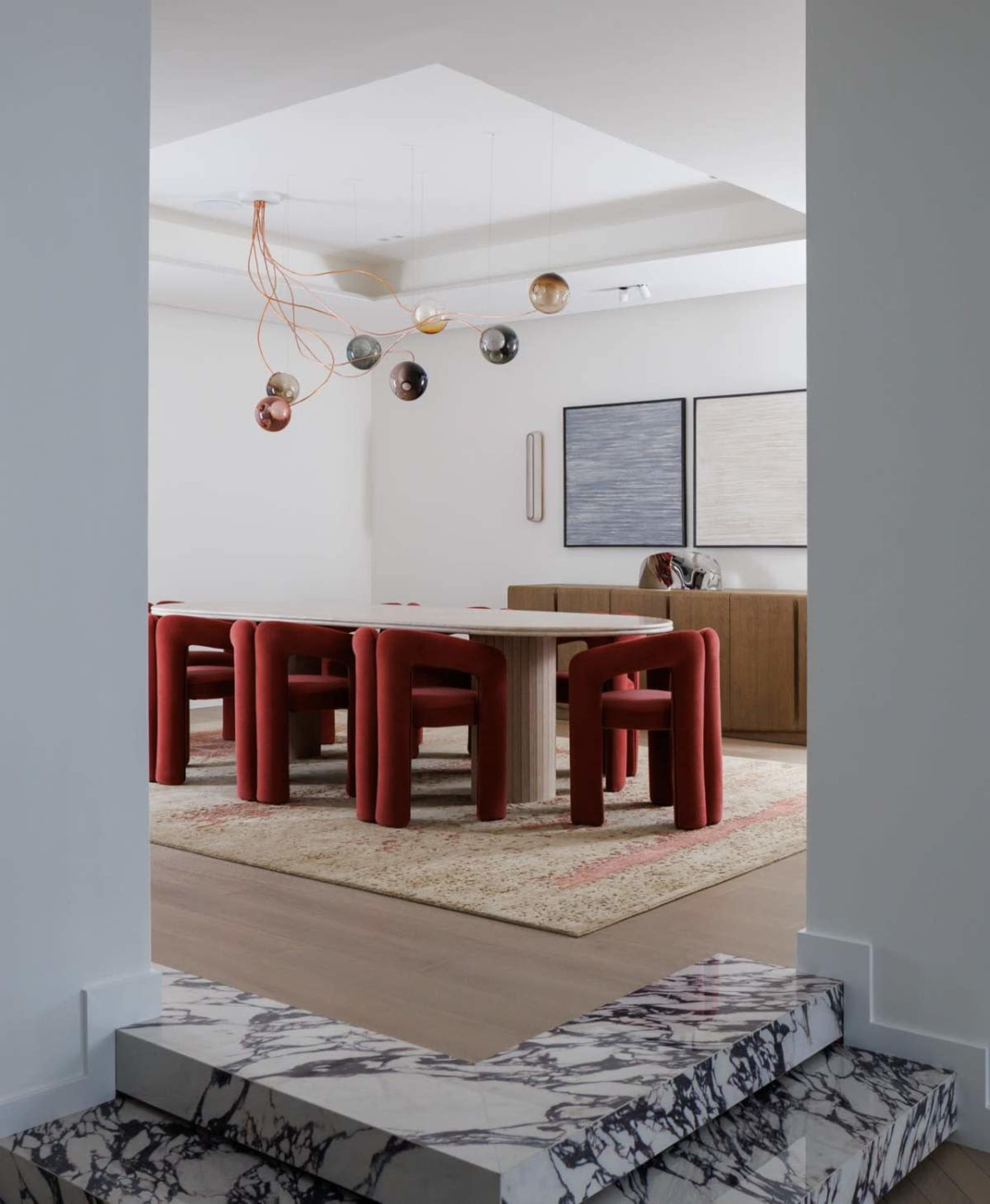 A dining area features a rectangular table surrounded by red upholstered chairs, with a pair of abstract artworks on the wall and a pendant light overhead.