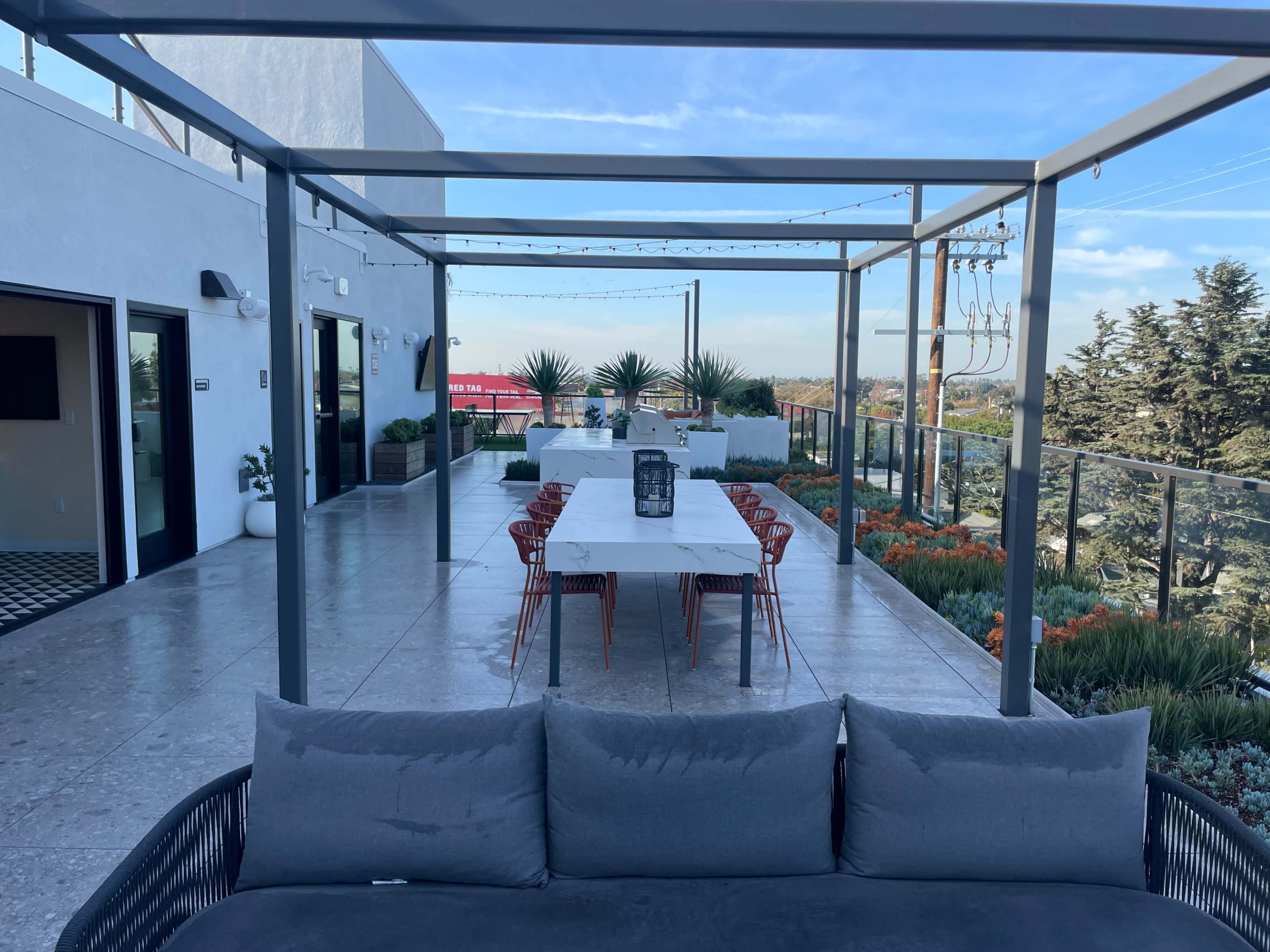 A spacious outdoor terrace with a long dining table and chairs under a metal pergola, surrounded by greenery and decorative plants.