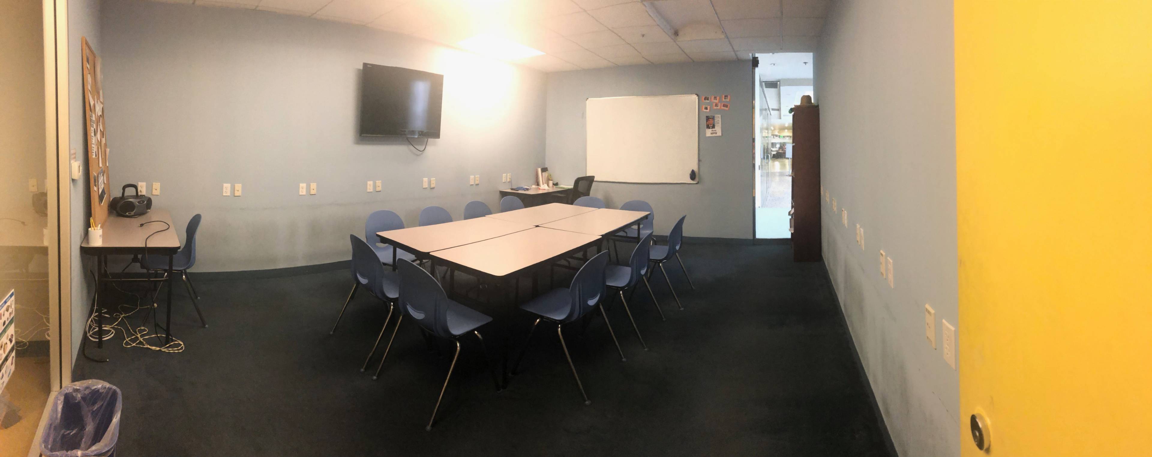 A spacious classroom with multiple tables arranged in a U-shape, blue chairs around them, a television mounted on the wall, and a whiteboard at one end.