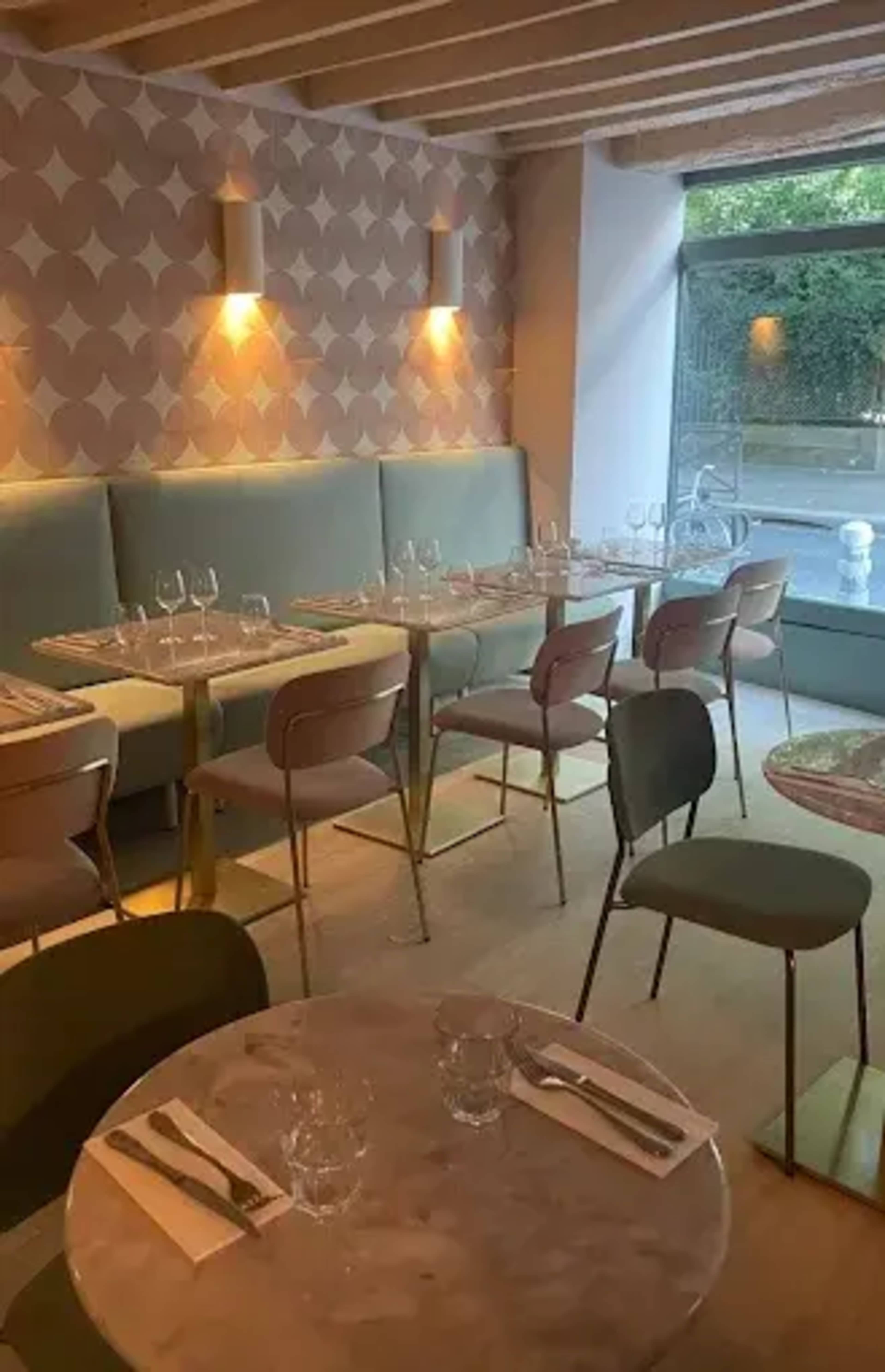The image shows the interior of a restaurant featuring light-colored table settings, a patterned wall, and a mix of modern seating arrangements.