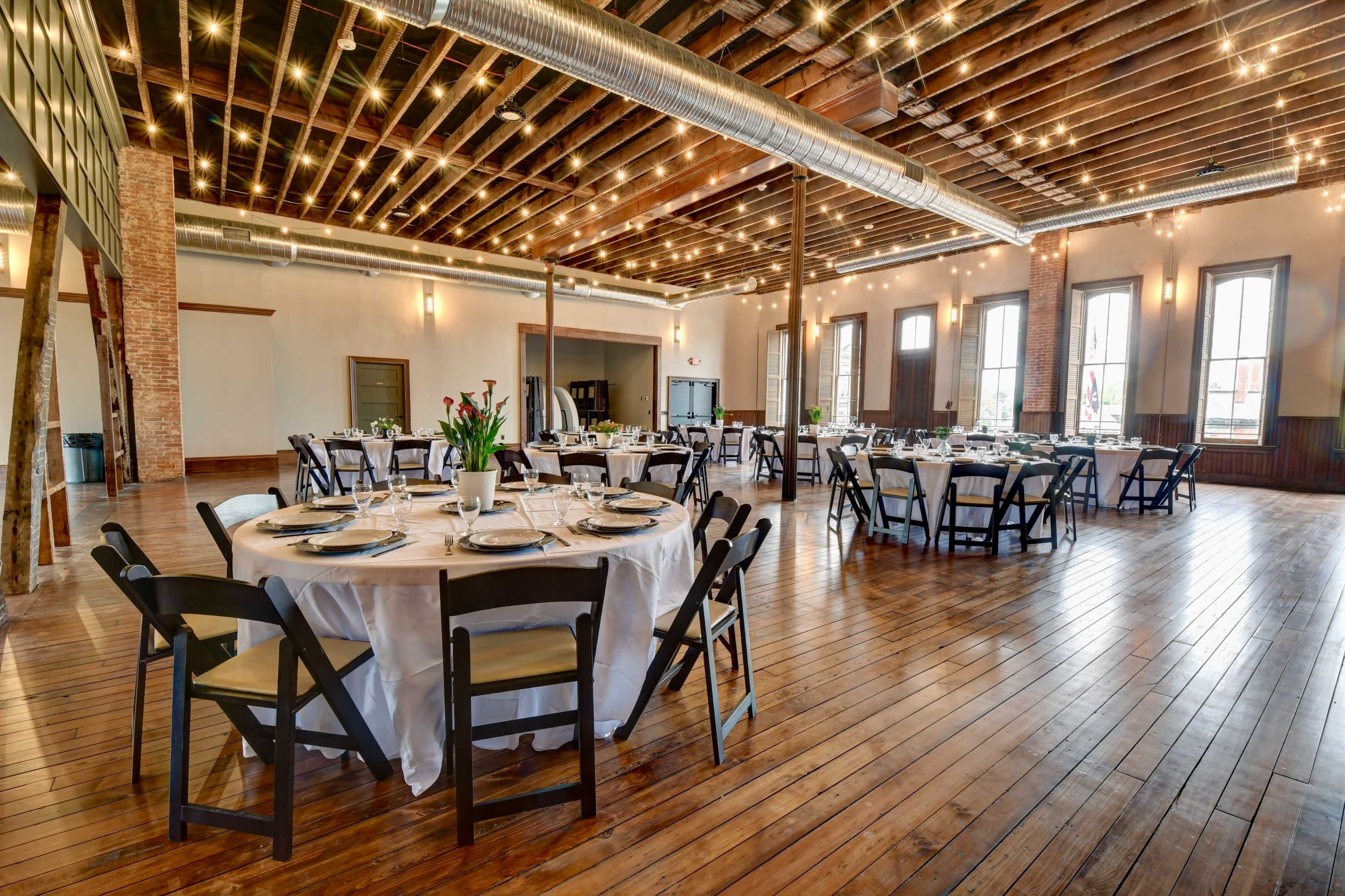 An elegant banquet hall with round tables set for dining, featuring wooden floors, exposed beams, and string lights overhead.