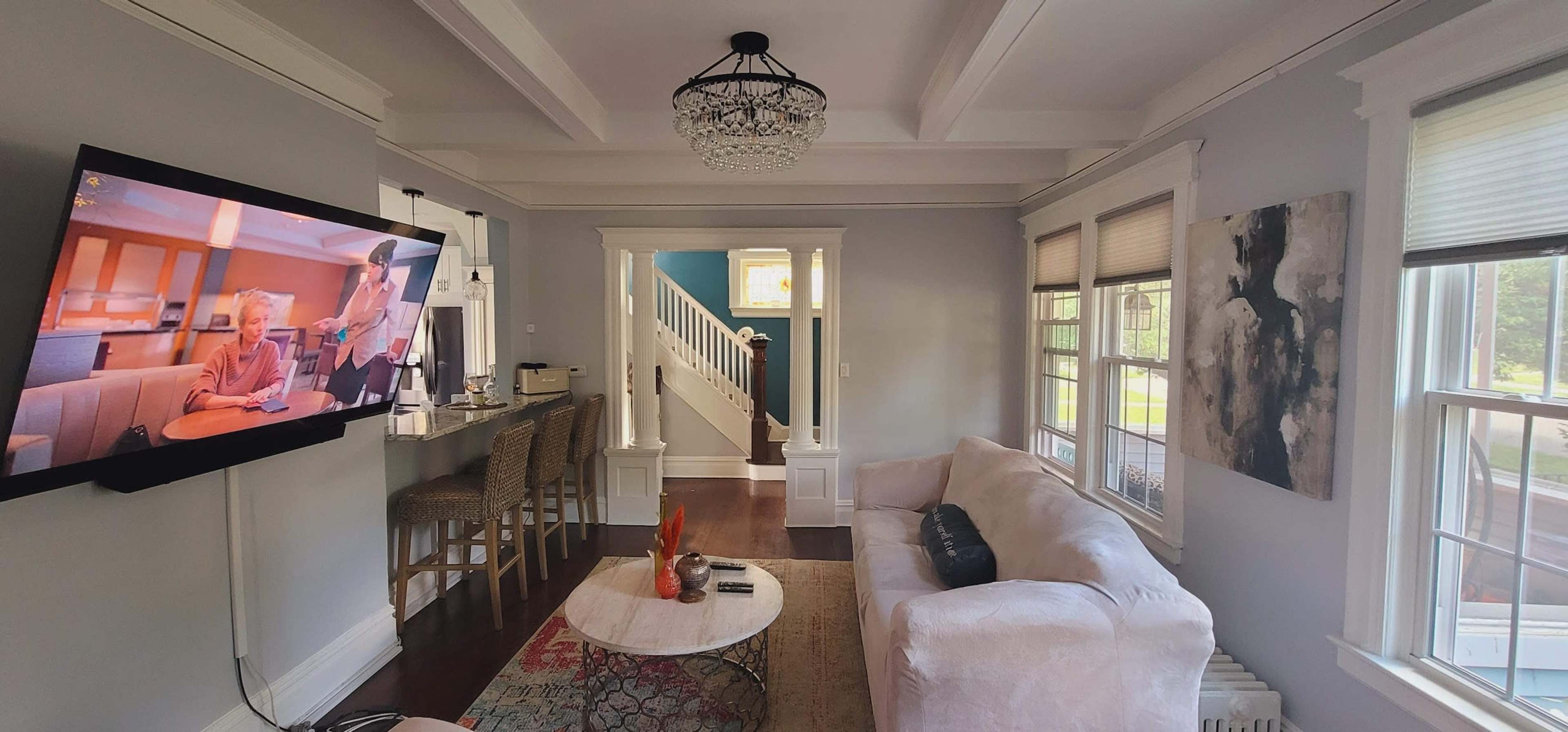 A living room with a large television mounted on the wall, a light-colored sofa, and a staircase visible in the background.