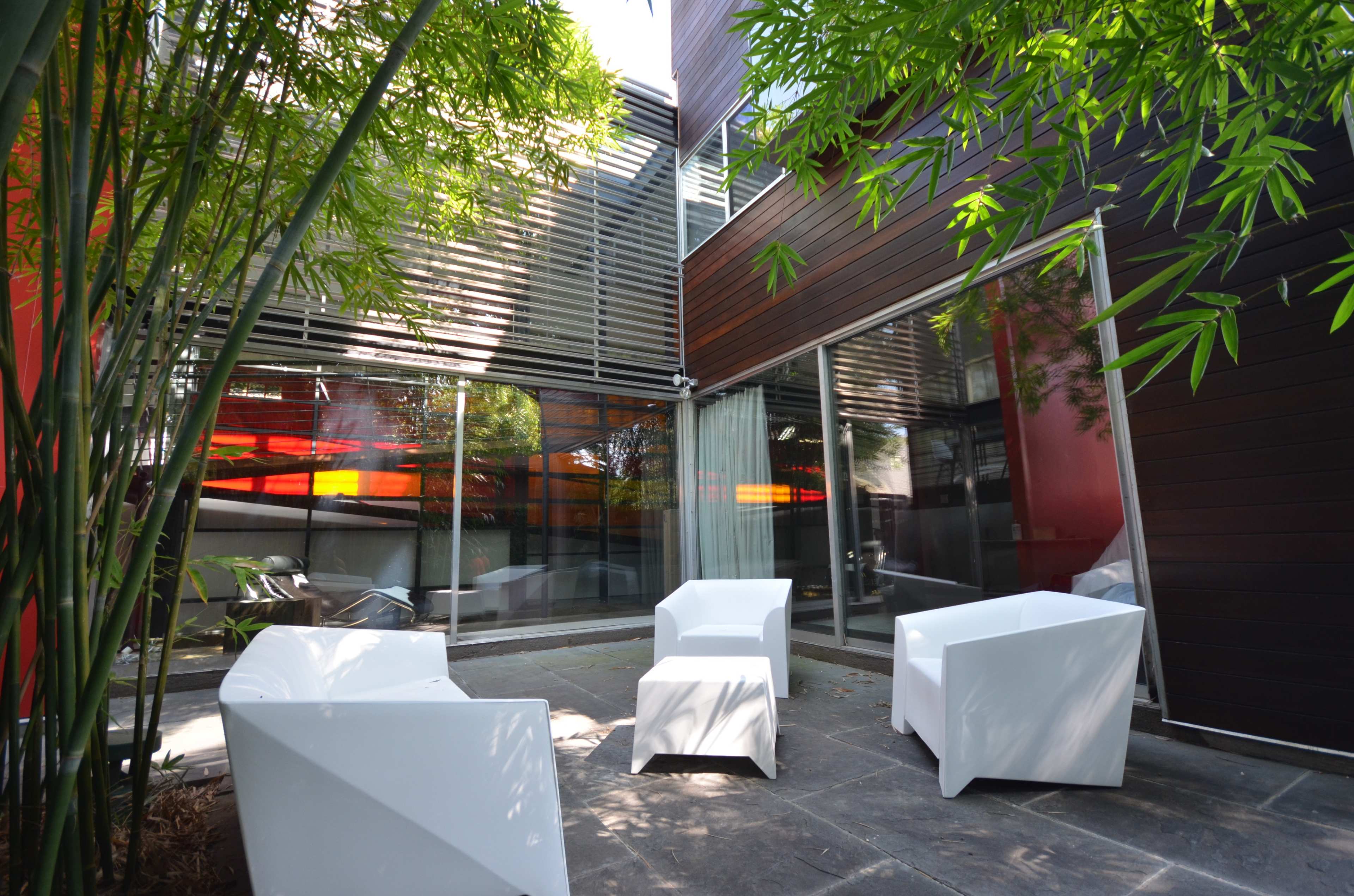 A modern outdoor seating area features white chairs and a stone patio surrounded by bamboo and large glass windows.