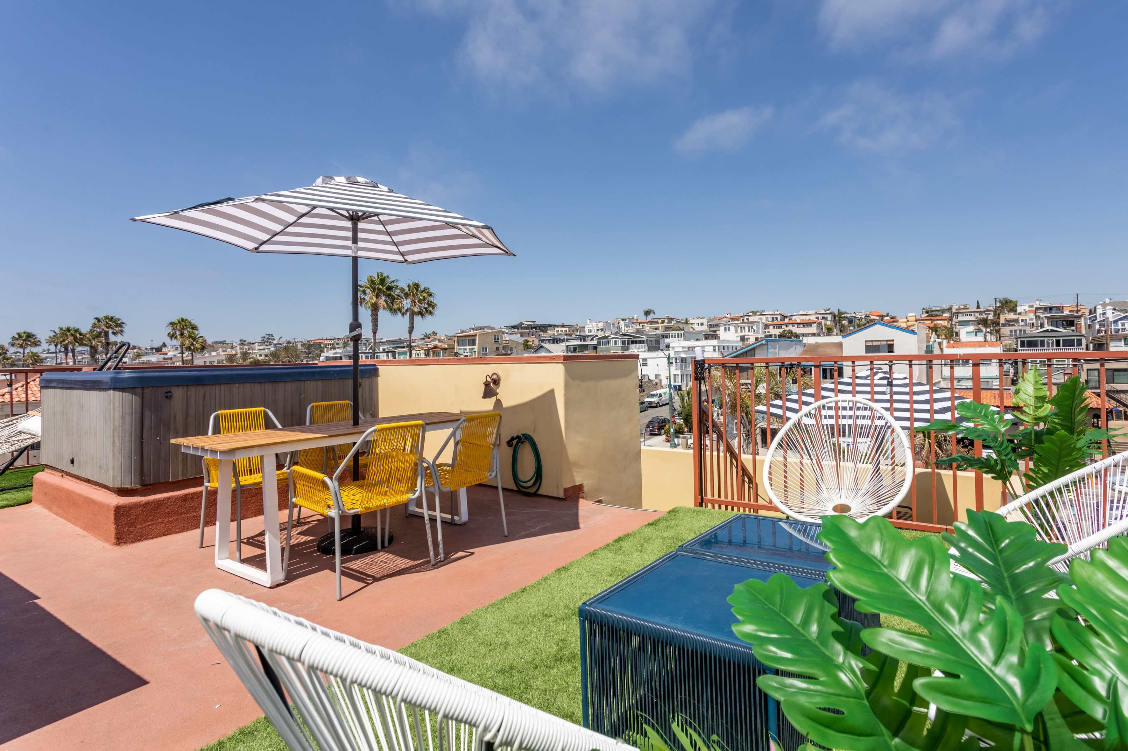 A rooftop patio features a table with yellow chairs, a striped umbrella, a hot tub, and views of nearby buildings and palm trees.