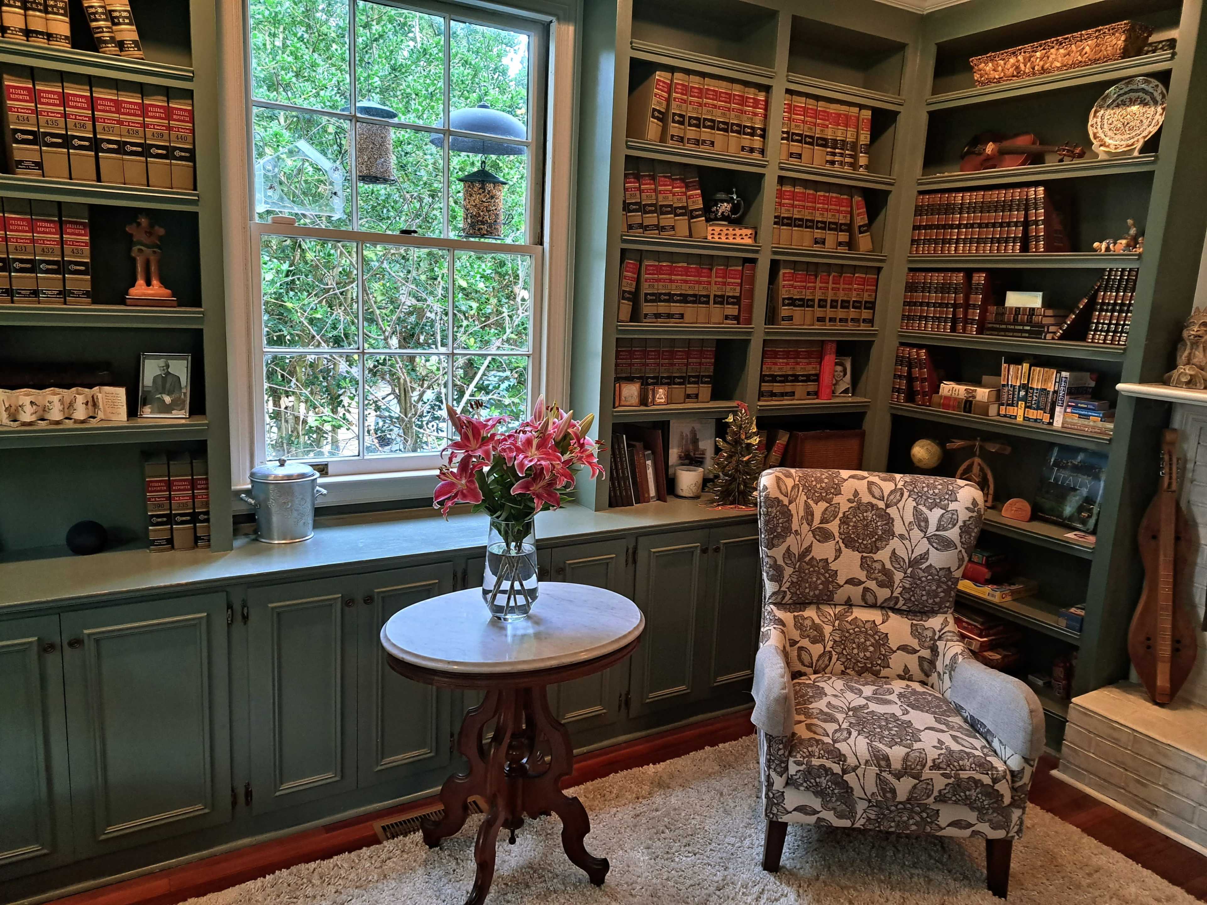 A cozy reading nook features a patterned armchair beside a round table, with bookshelves filled with law books and a window letting in natural light.