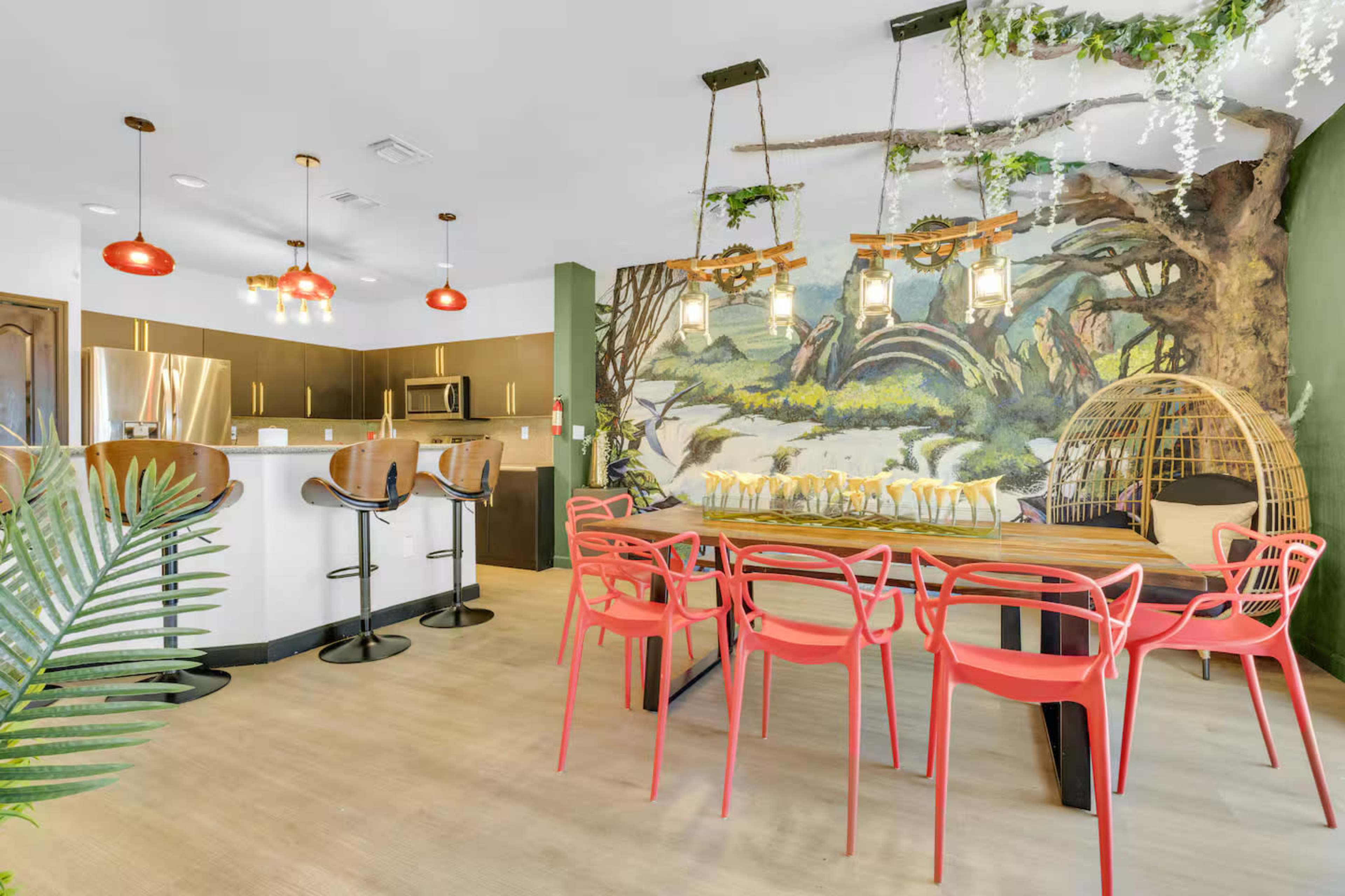 The image shows a modern kitchen and dining area featuring a large wooden table with red chairs, bar seating at a kitchen island, and a mural of a natural landscape on the wall.