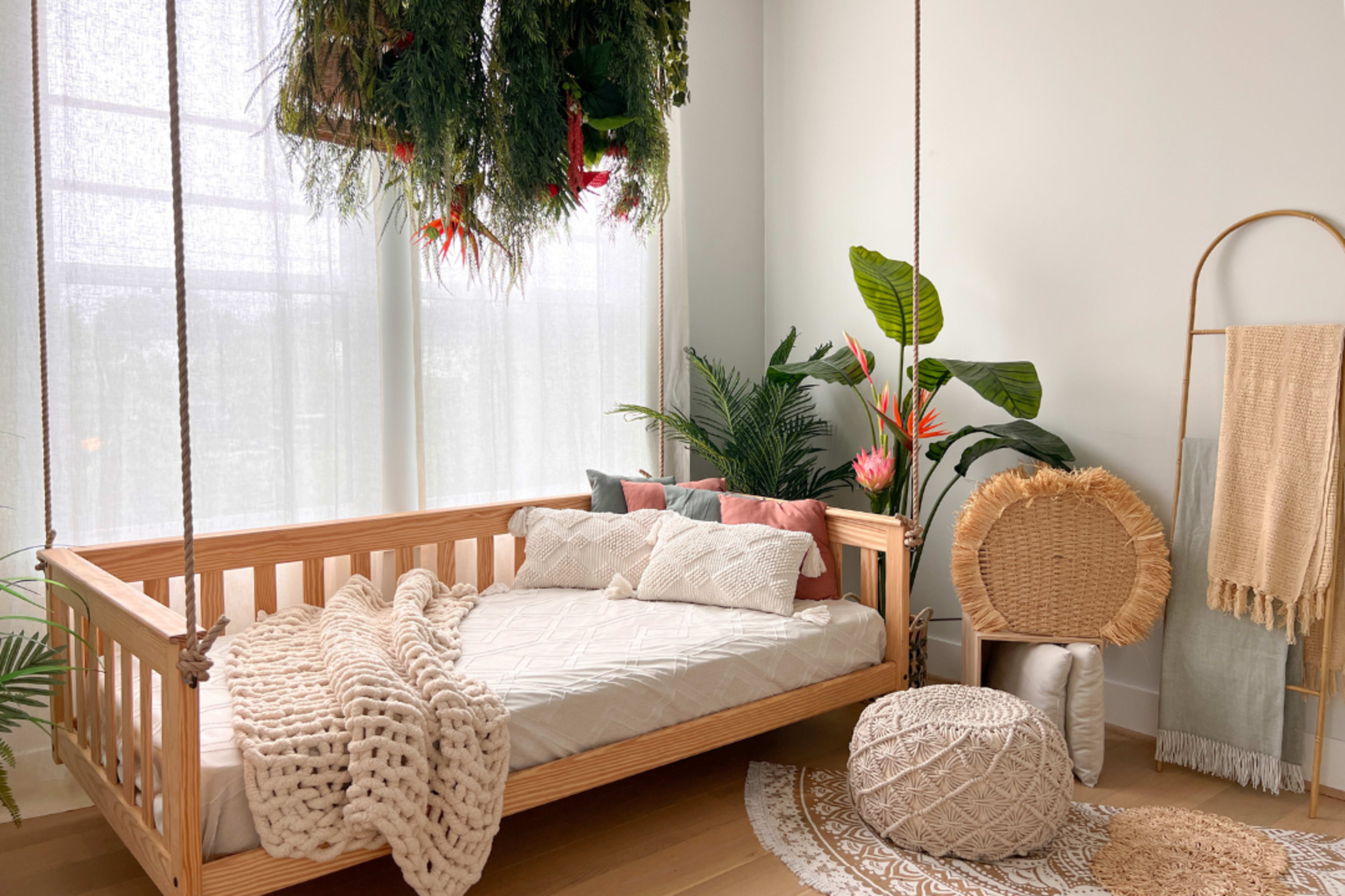 The image shows a light-filled room with a wooden swing bed adorned with pillows and a crochet blanket, surrounded by various plants and decor elements.