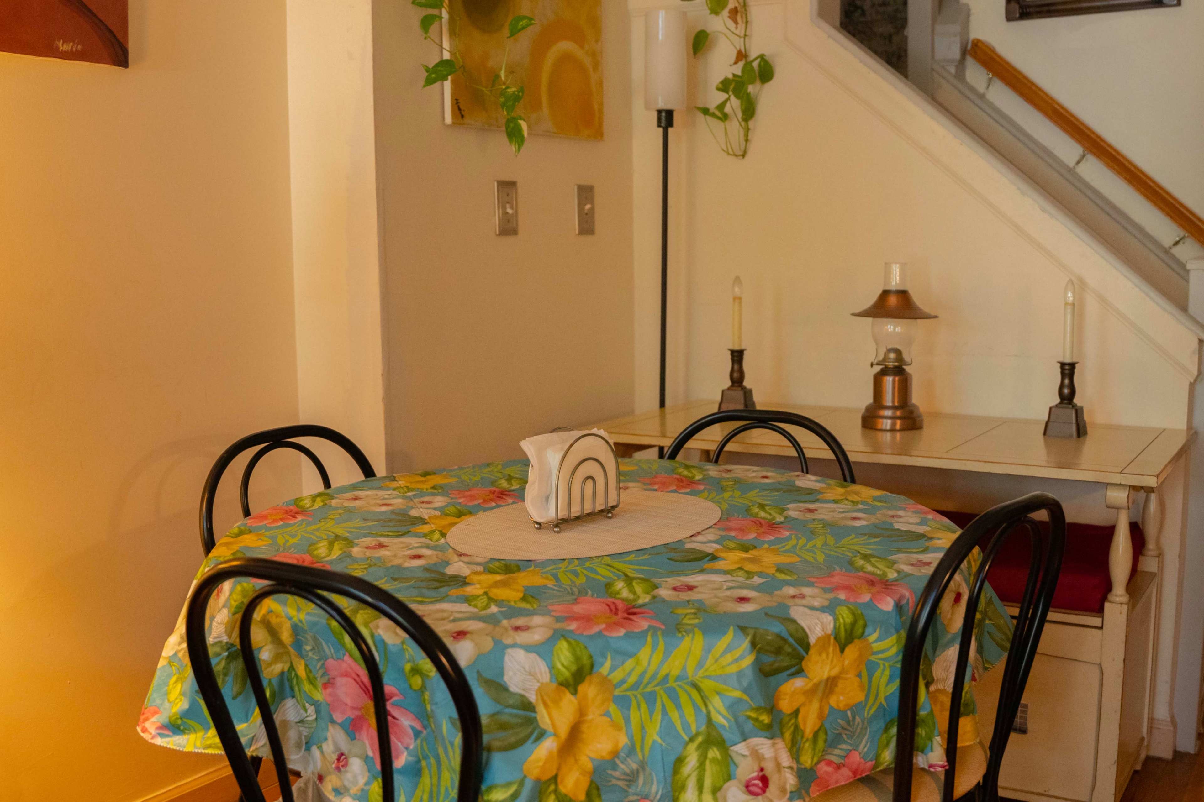 The image shows a small dining area with a round table covered in a floral tablecloth, surrounded by black chairs, and a lamp beside a staircase.