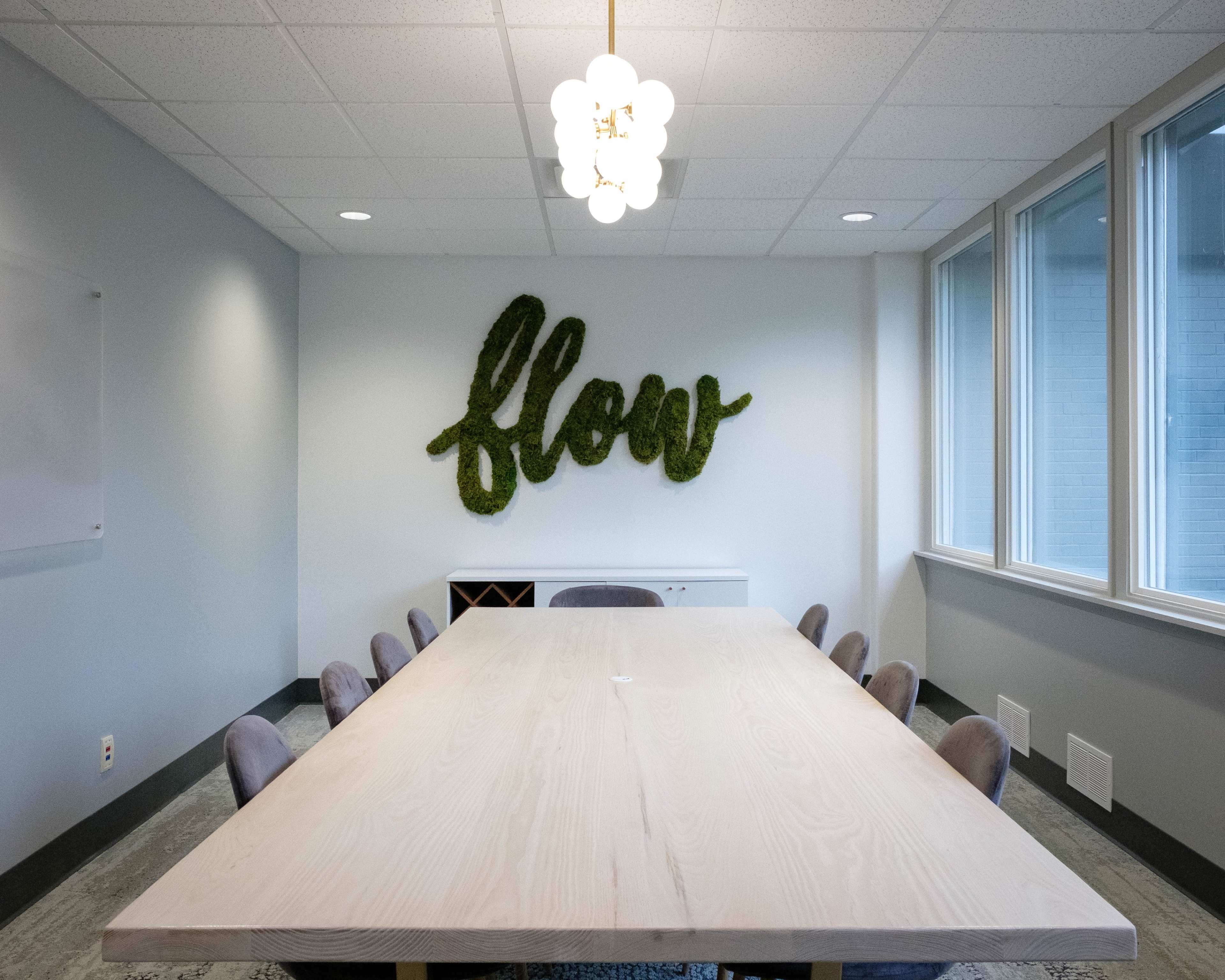 A long wooden table is set in a brightly lit conference room featuring a wall-mounted green "flow" sign made of foliage.