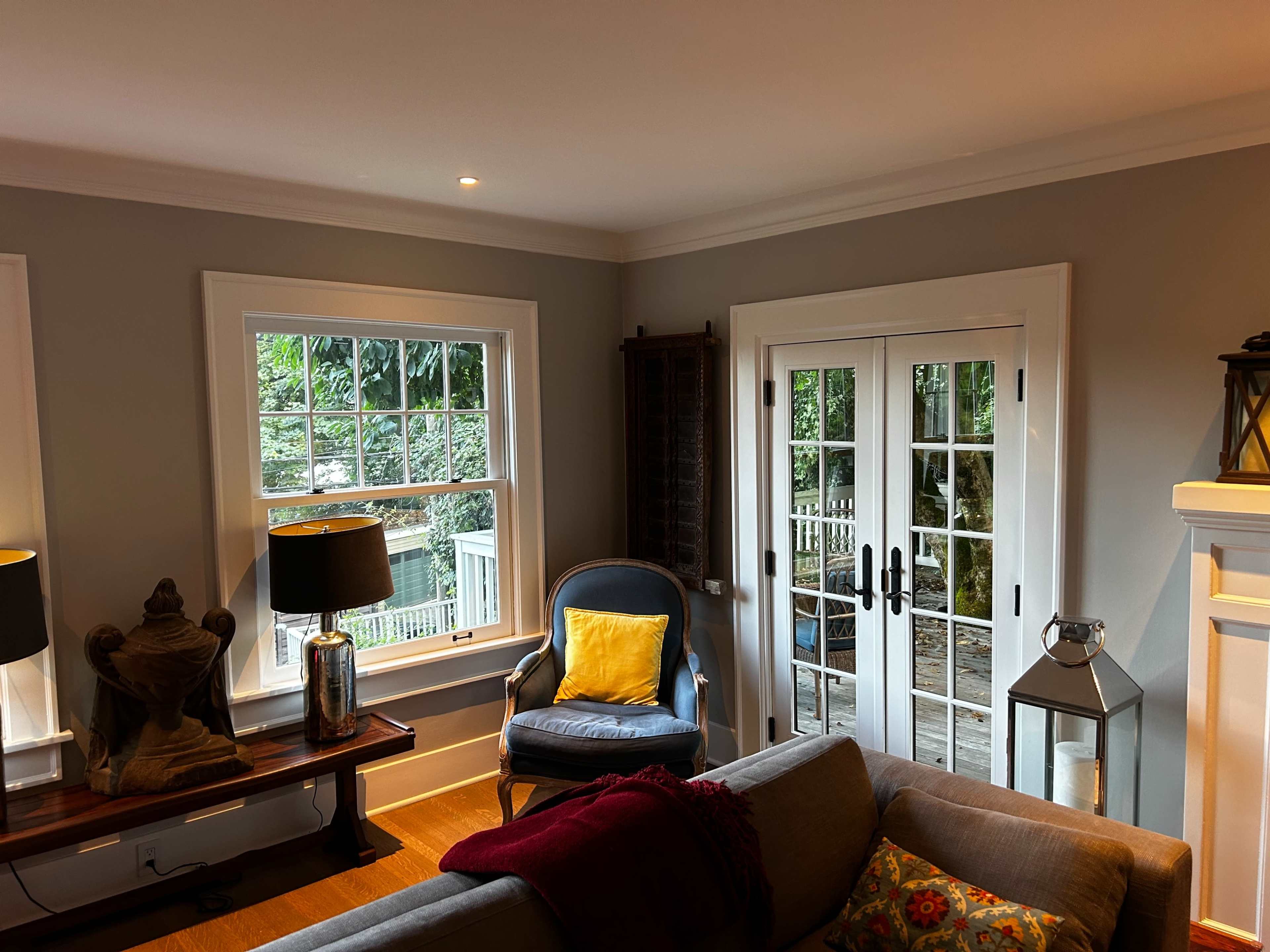 A cozy living room with large windows, a blue armchair, a wooden coffee table, and double French doors leading to an outdoor area.