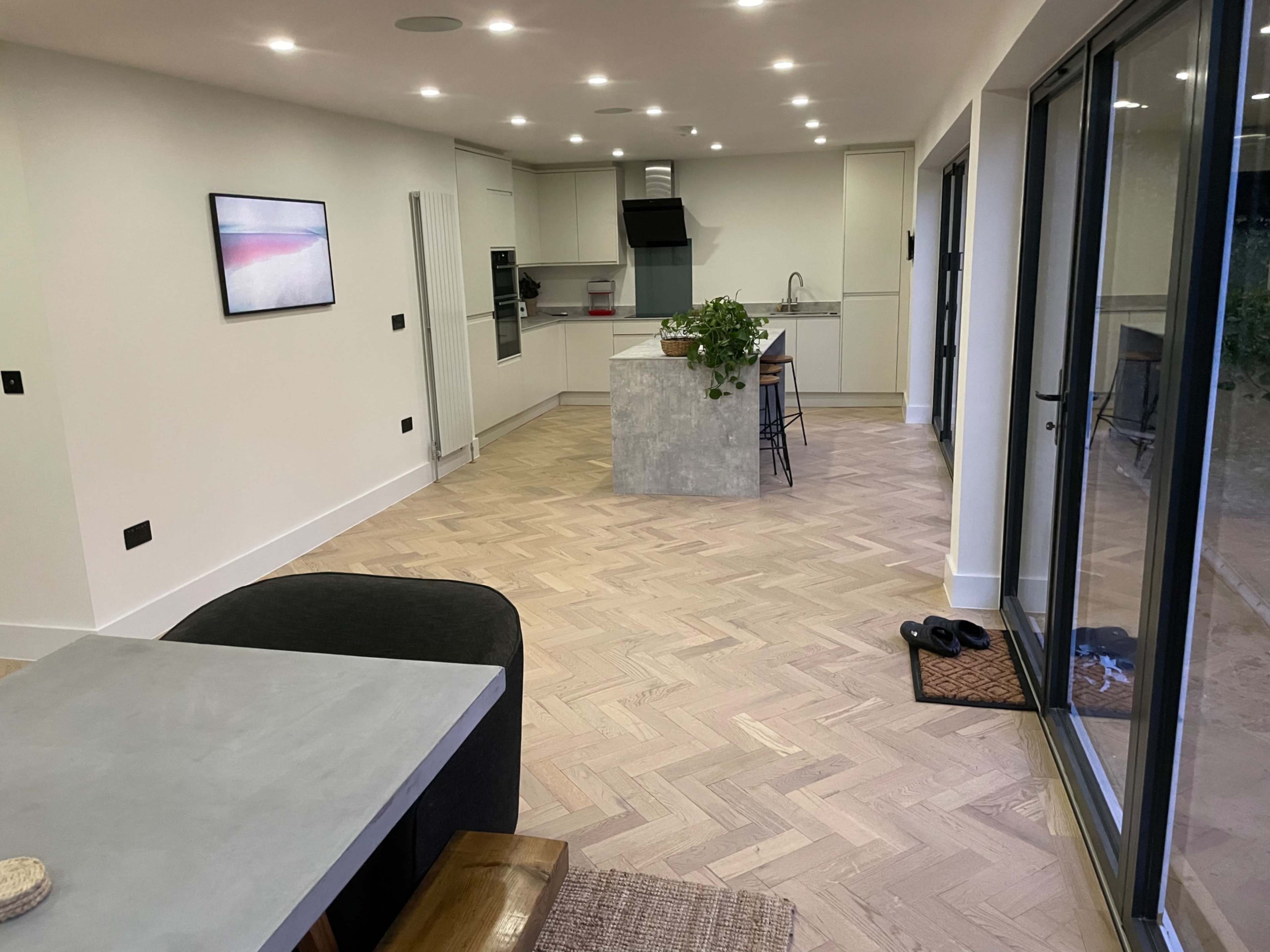 A modern kitchen and dining area with a light-colored herringbone wooden floor, a central island with a stone surface, and large windows allowing natural light.