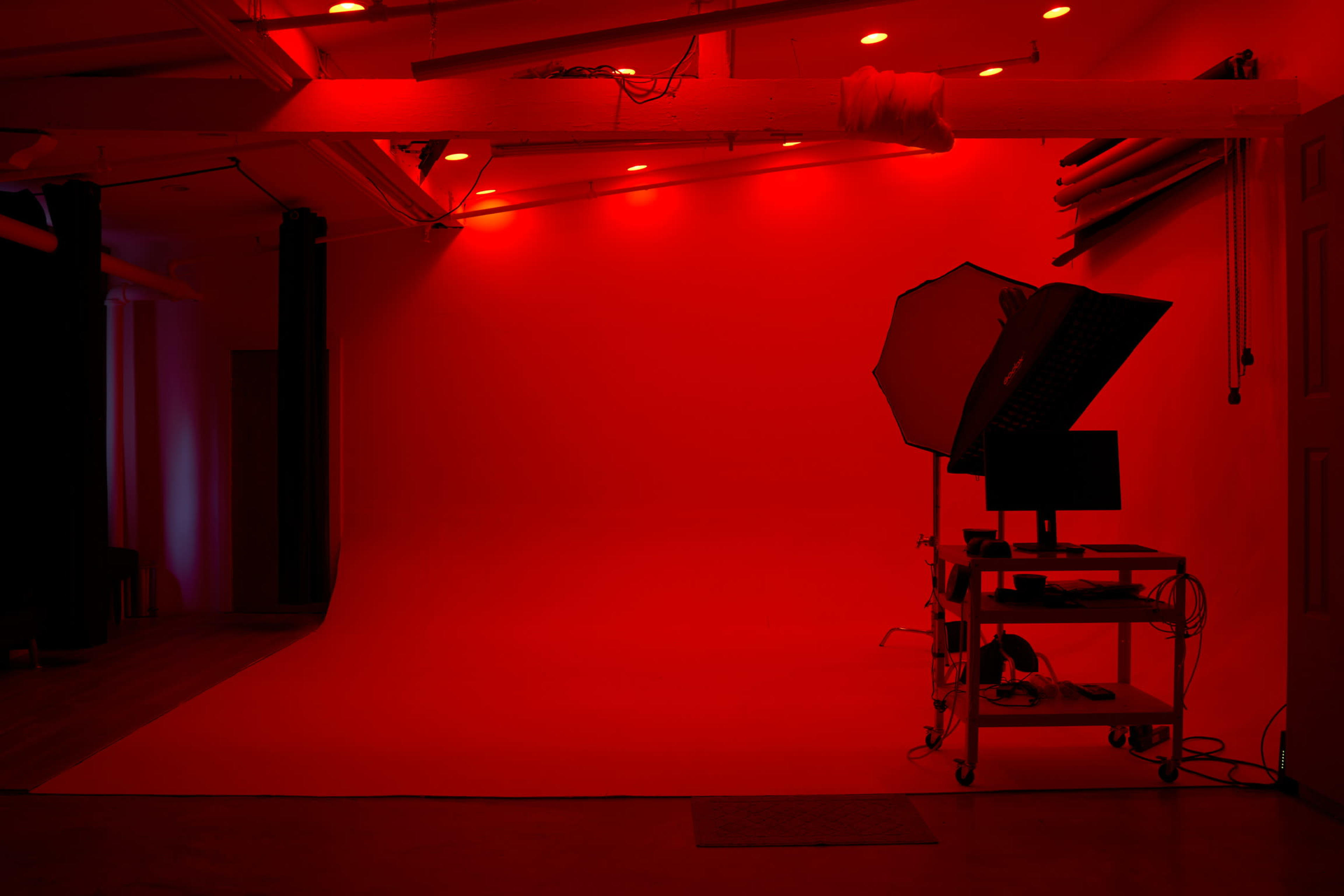 A photo studio is illuminated with red lights, featuring a seamless backdrop and a cart with a computer and lighting equipment.
