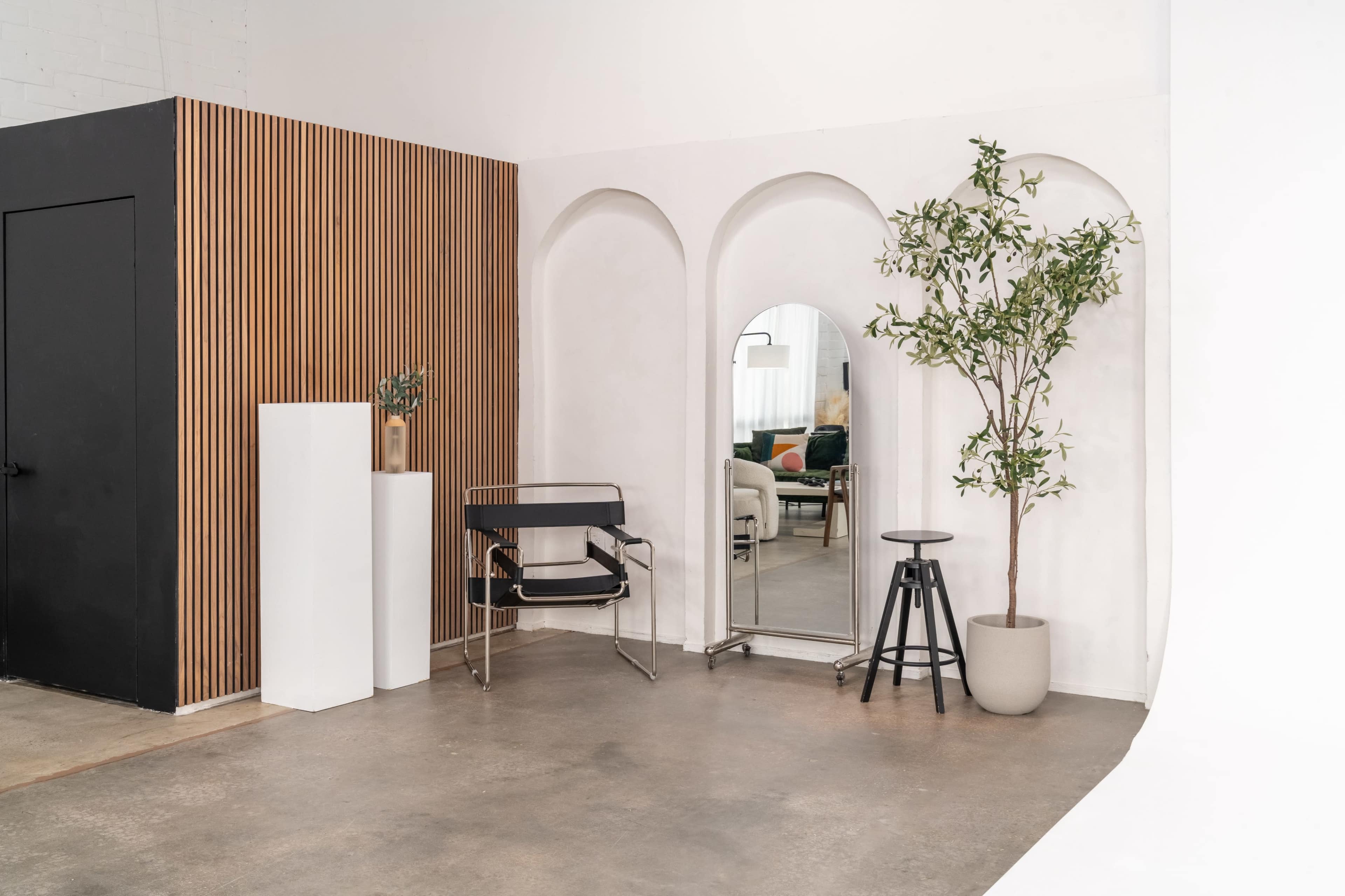 A modern interior space featuring arched wall openings, a large mirror, and minimalist furniture, including a chair and a stool, set against a backdrop of wood paneling and a potted plant.