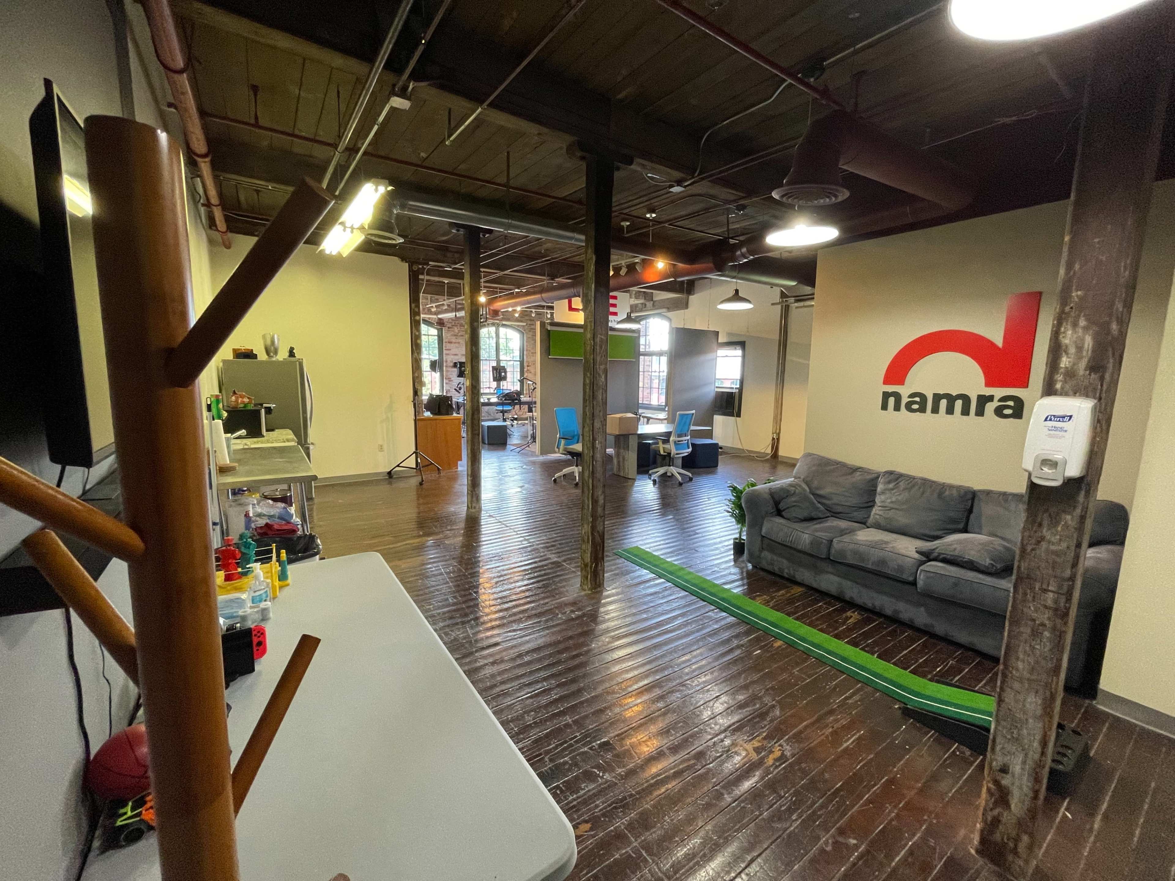 An office interior featuring a gray couch, a wooden coat rack, and a green track on the floor, with a large wall logo for "namra" in red.
