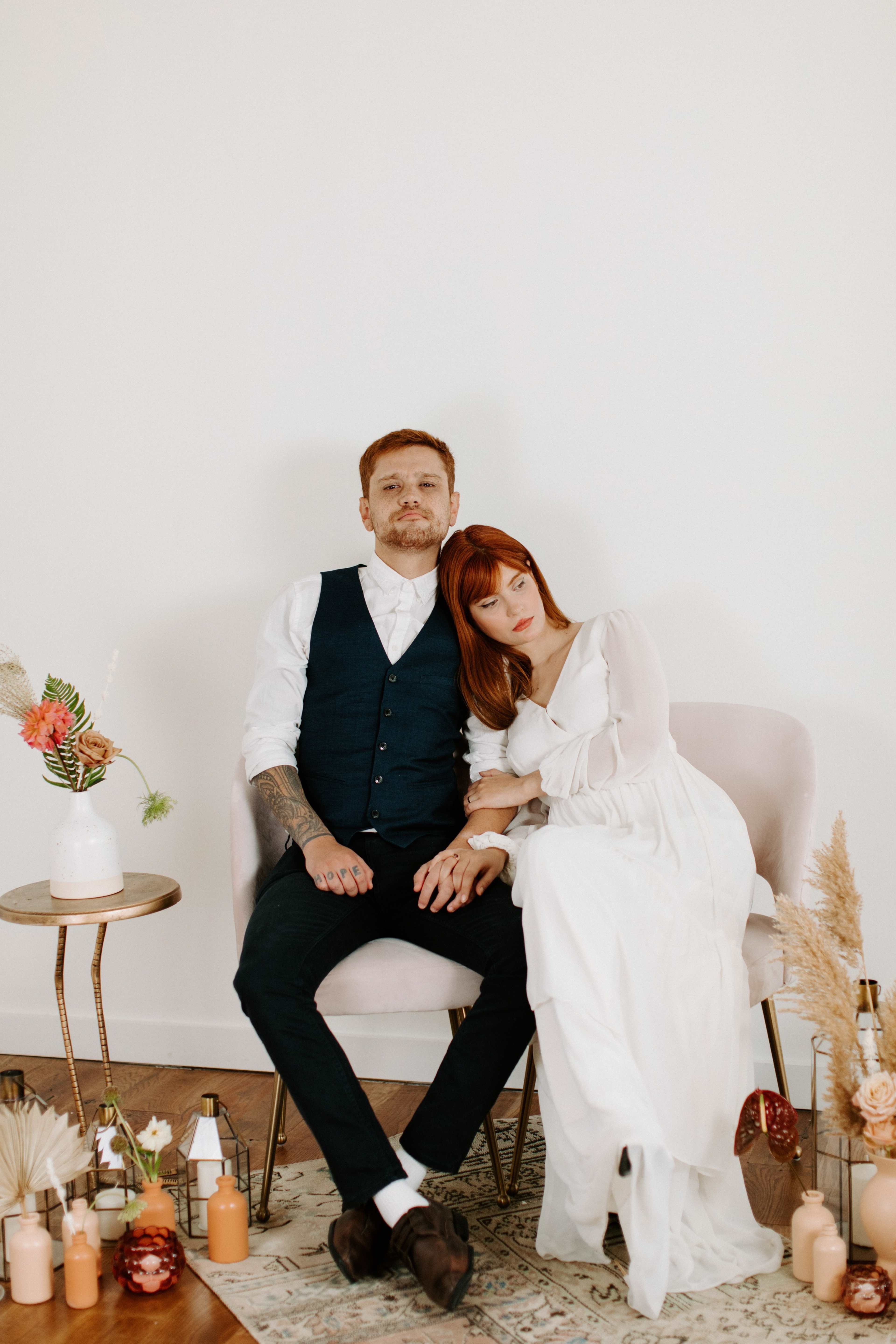 A couple sits closely together on a pink chair, with the woman resting her head on the man's shoulder in a softly decorated indoor setting.