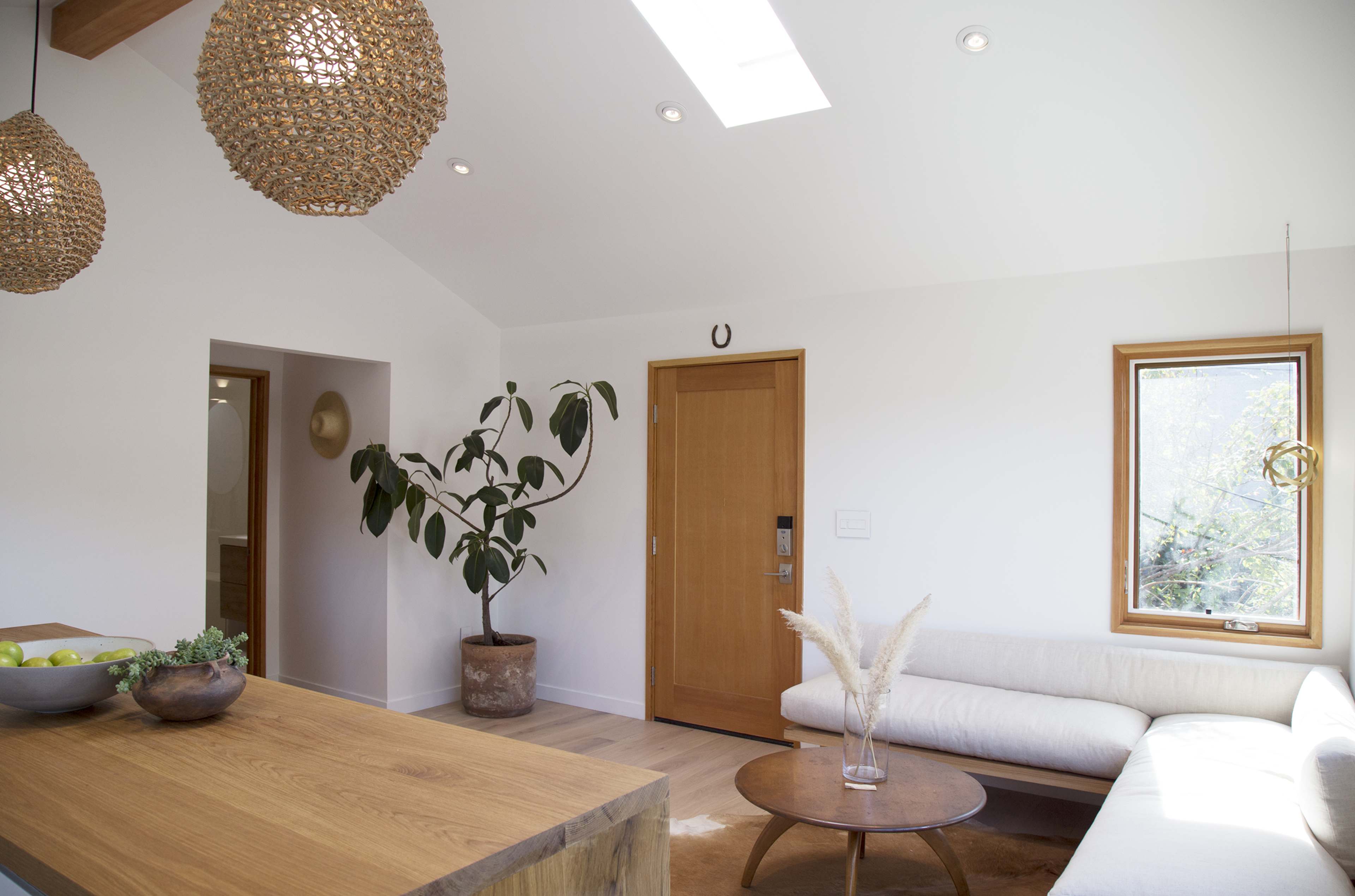 A modern living space with a wooden table, a plant in a pot, a light-colored sofa, and natural light coming through a skylight and window.
