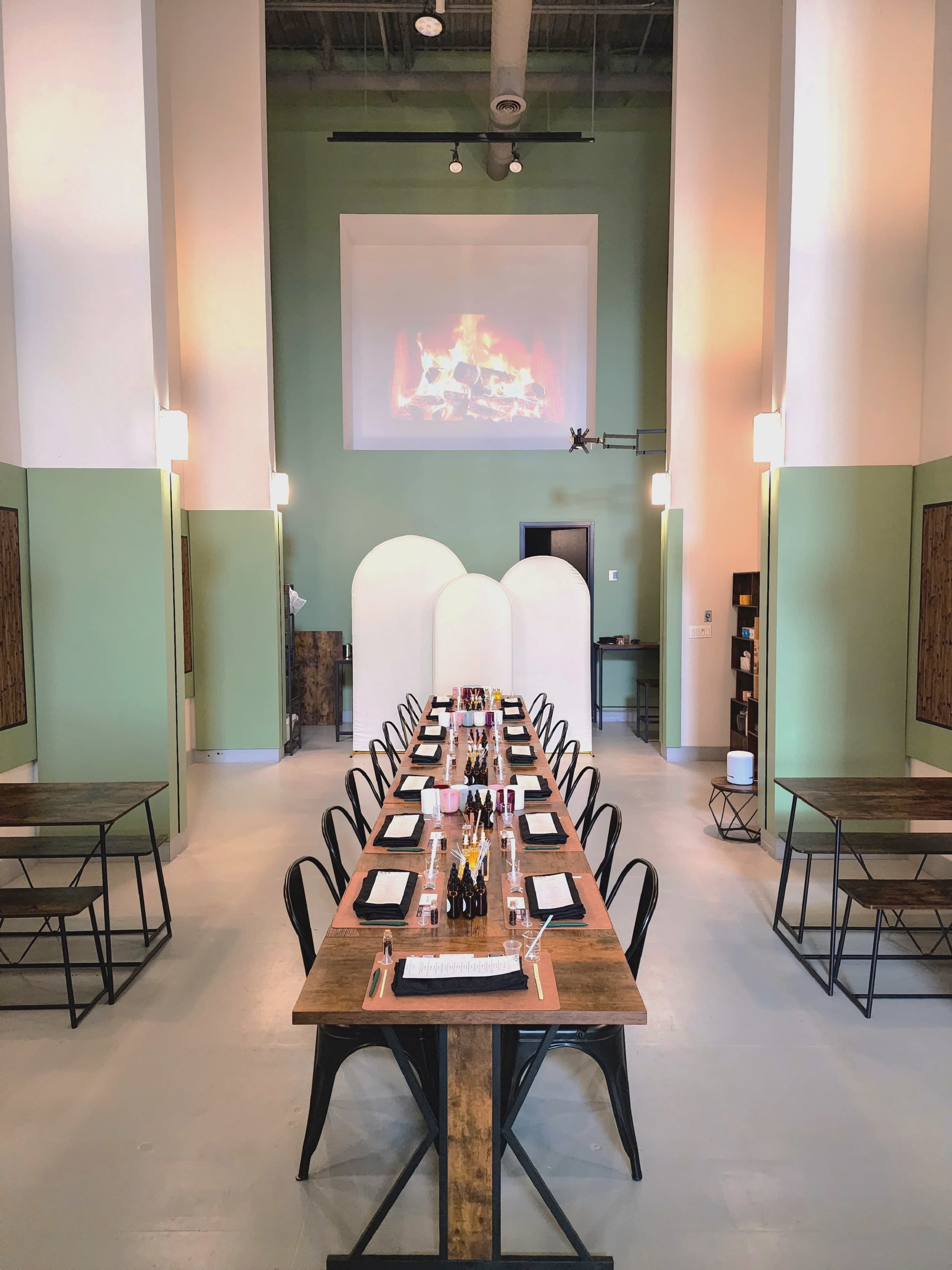 A long dining table is set in a spacious room with green walls and a projection of a fireplace on the wall.