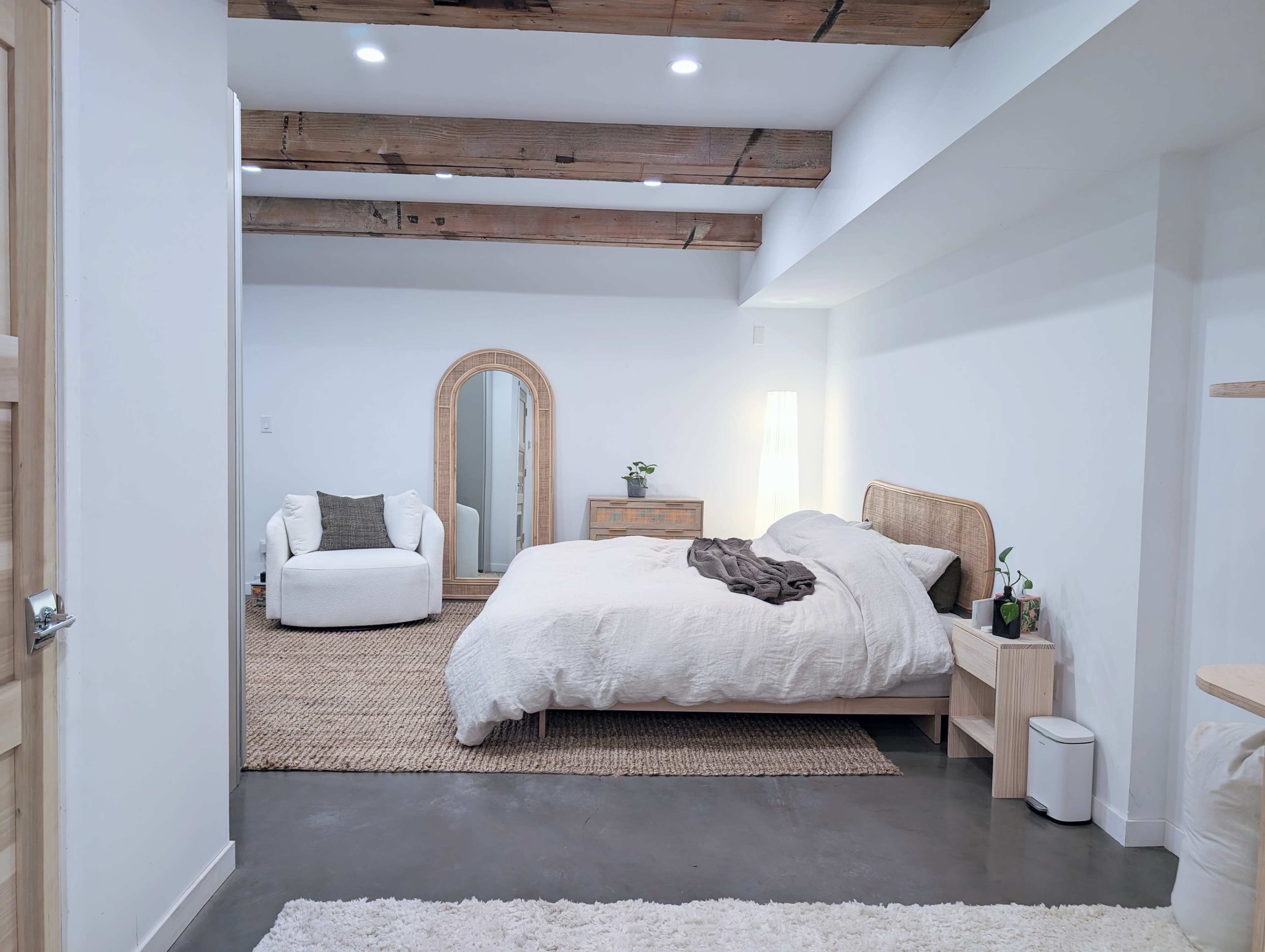 A minimalist bedroom with a bed, a mirror, an armchair, and wooden beams on the ceiling.