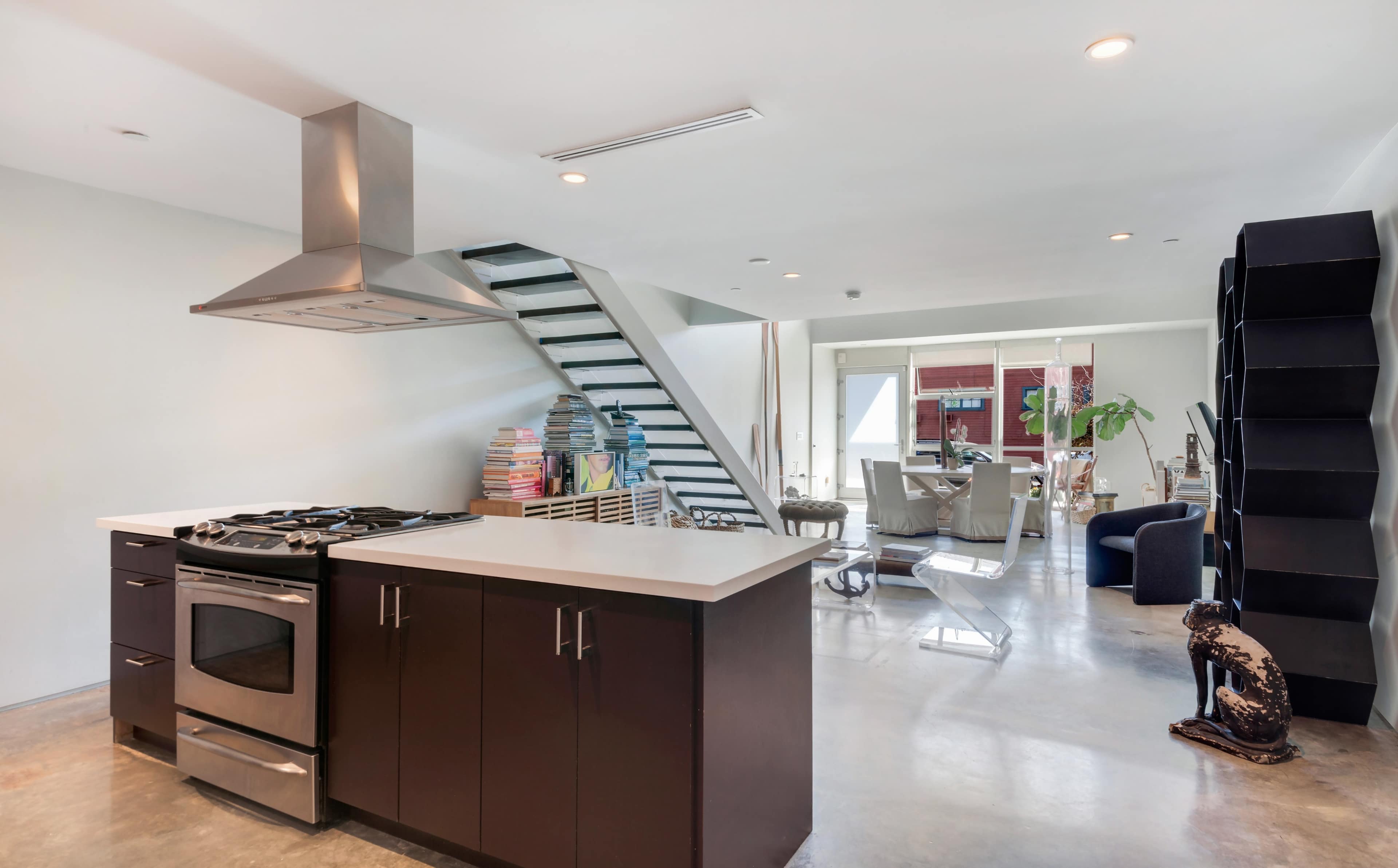 The image shows a modern kitchen with a central island, stainless steel appliances, and an open layout leading to a living area and staircase.