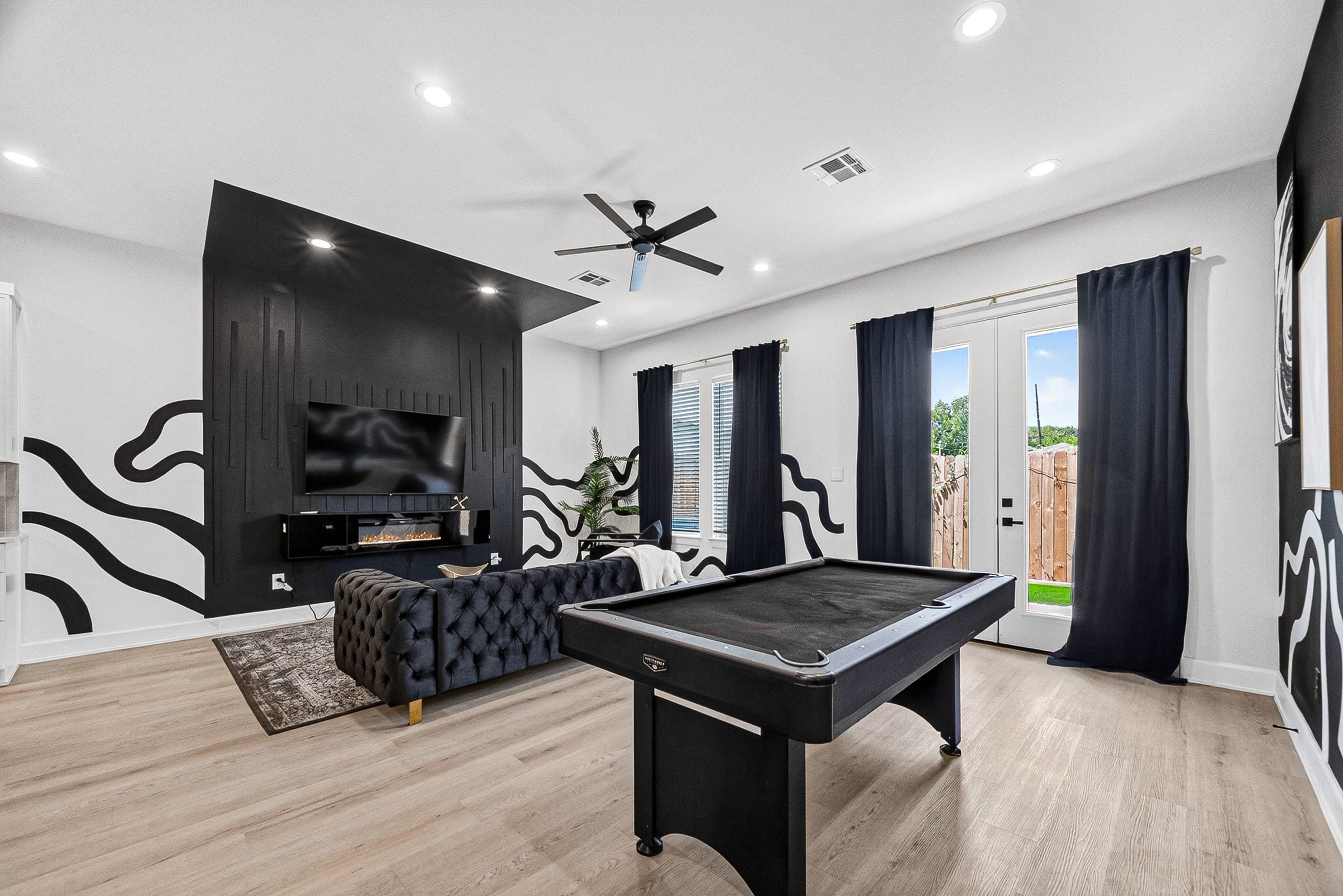 The image shows a modern living room featuring a black pool table, a dark sectional couch, and a large television mounted on a black accent wall.