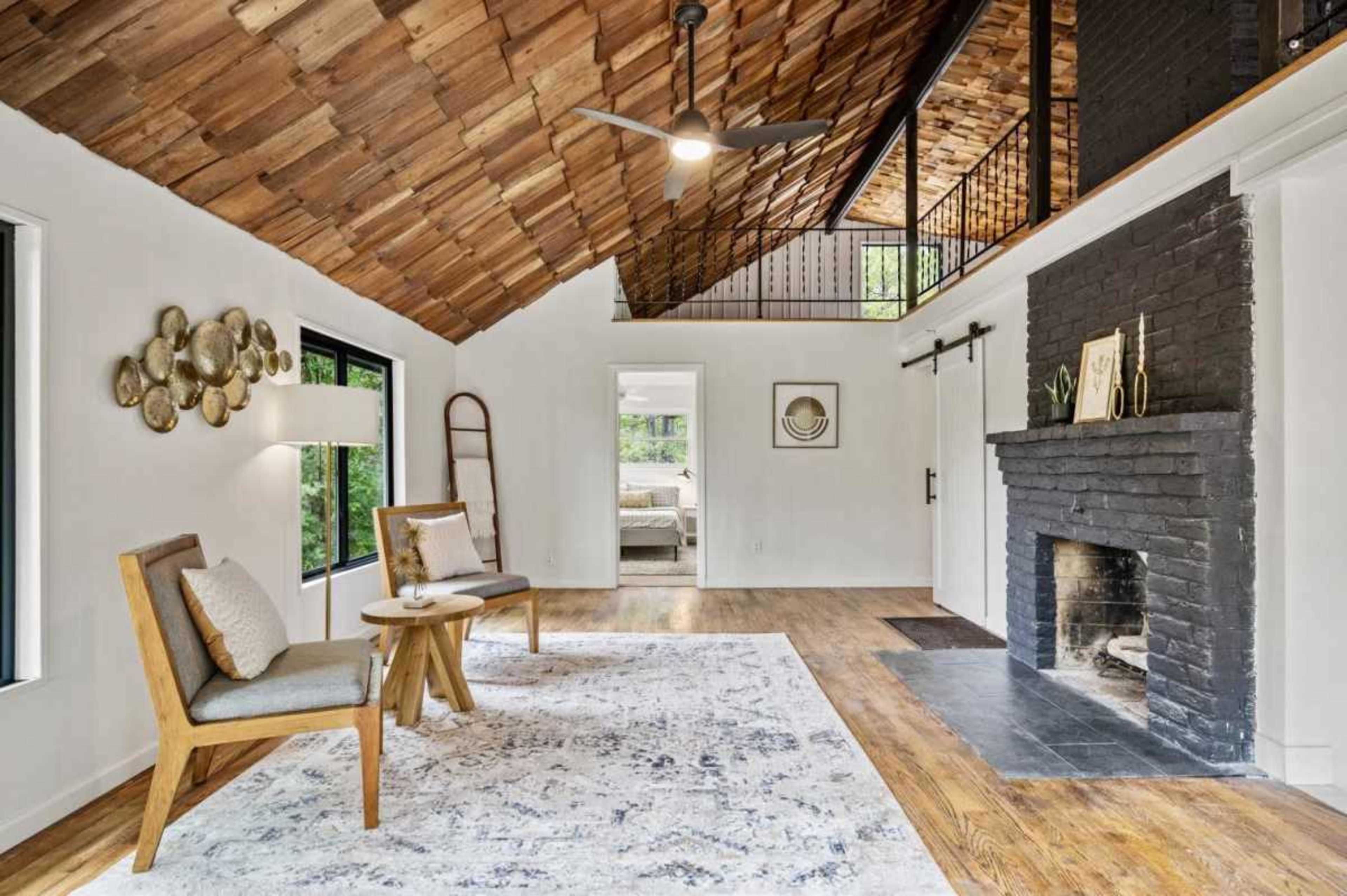 The image depicts a modern interior living space featuring a wooden ceiling, a black brick fireplace, two chairs, and a patterned rug on a hardwood floor.