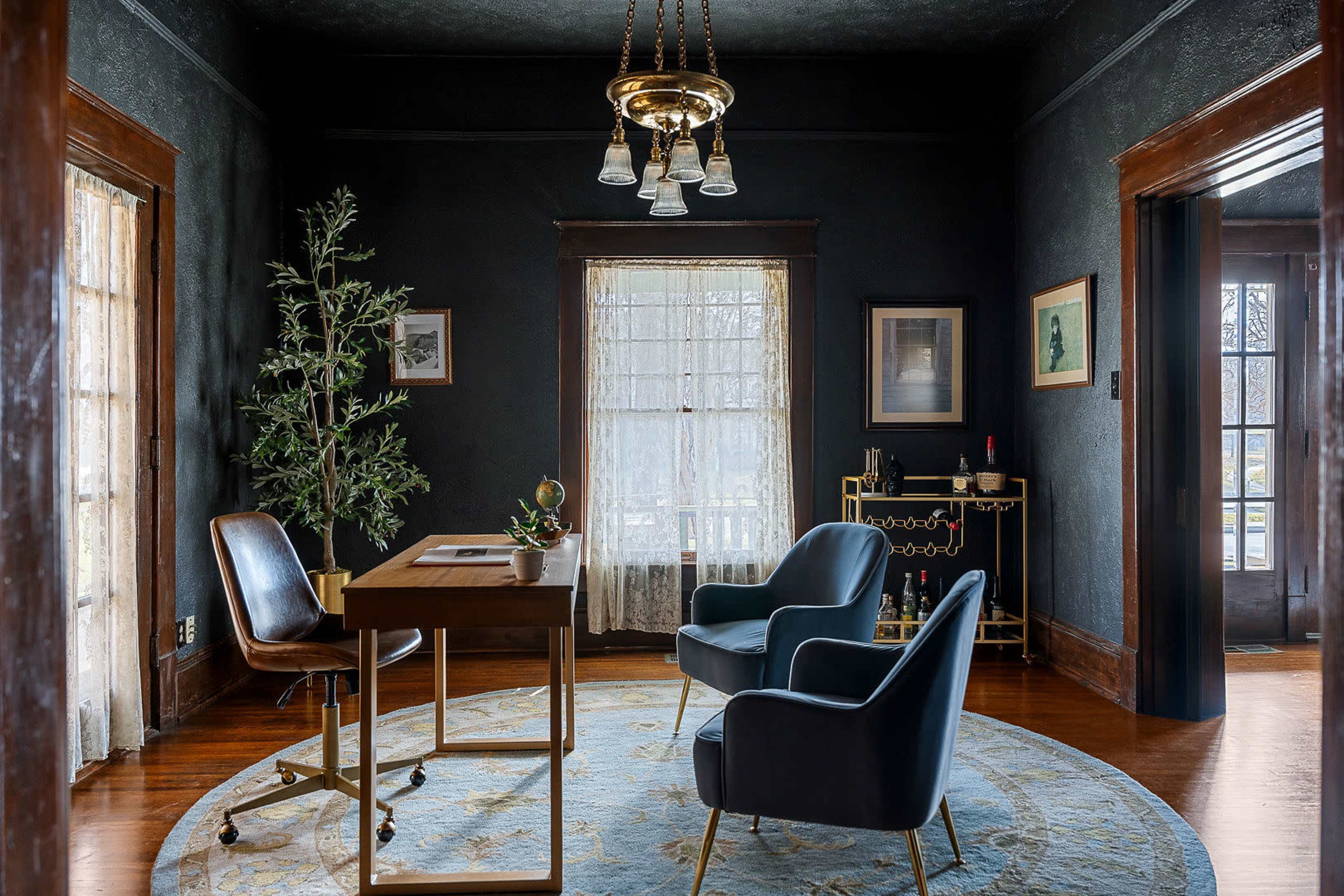 A modern office space features a wooden desk, two blue armchairs, a round area rug, and decorative plants against dark walls and large windows.