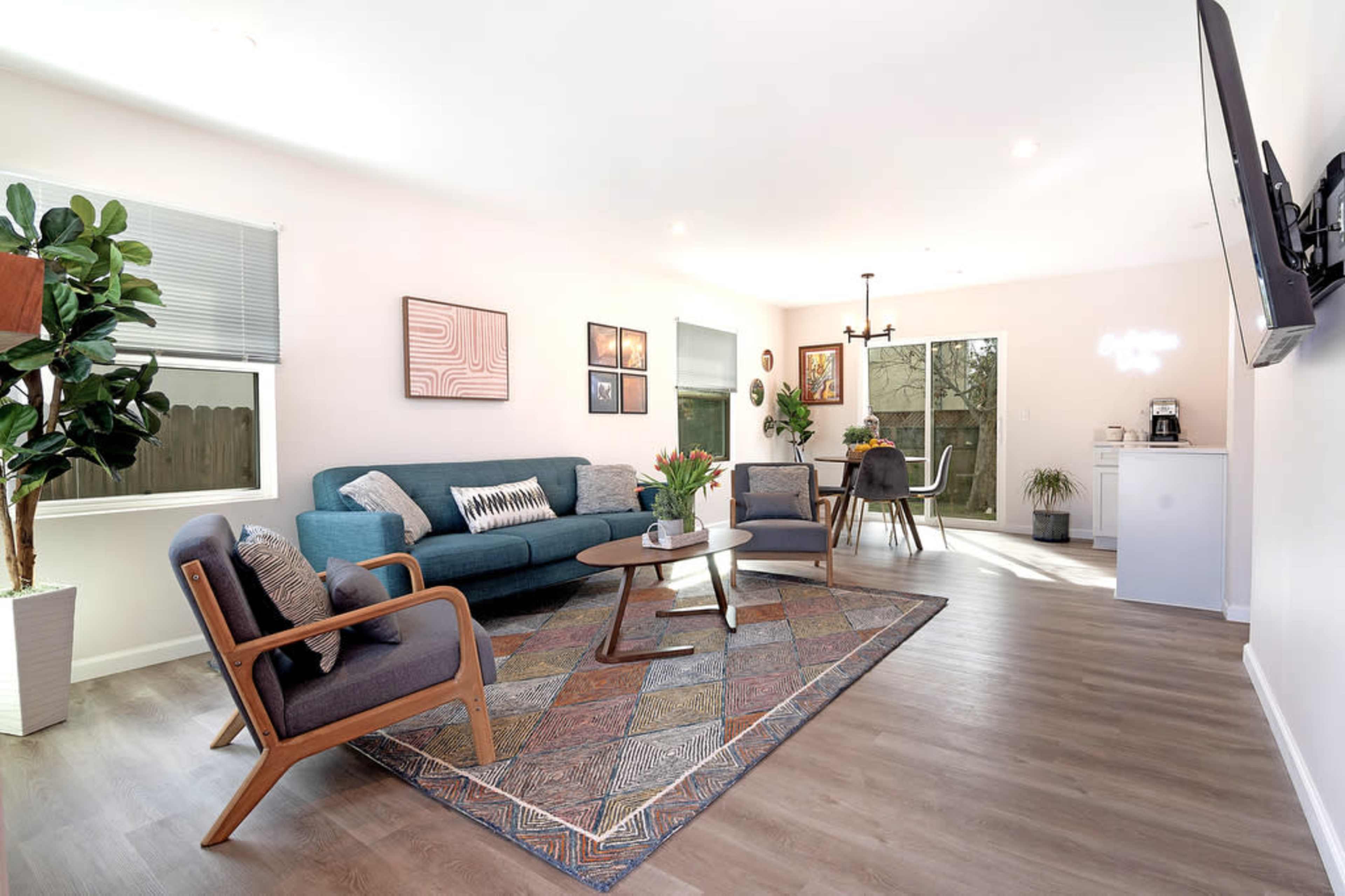 A modern living room features a blue sofa, two wooden armchairs, a coffee table, and a large area rug, with a dining area visible in the background.