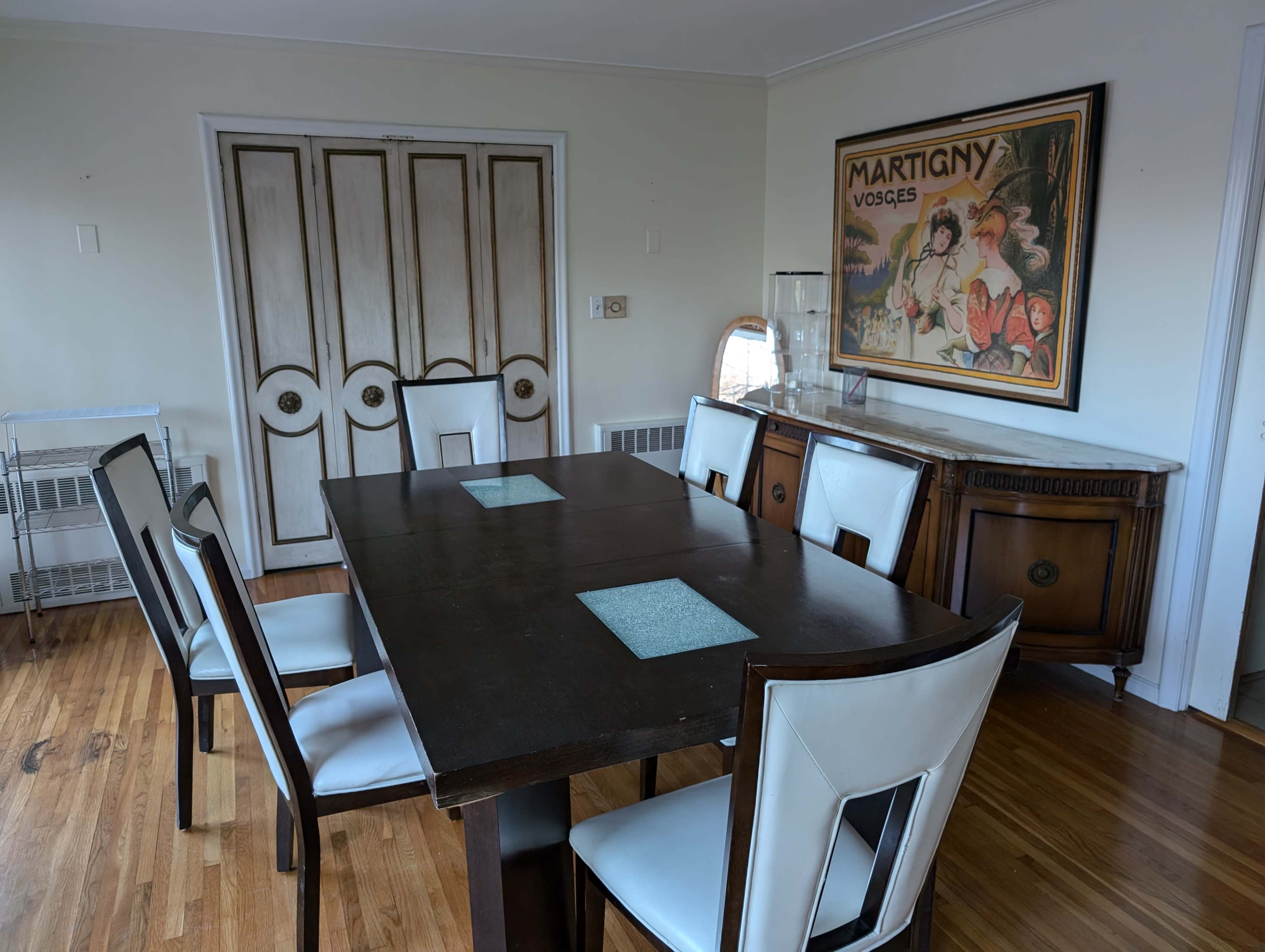 A dining room features a wooden table surrounded by white chairs, with a poster on the wall and a sideboard displaying glassware.