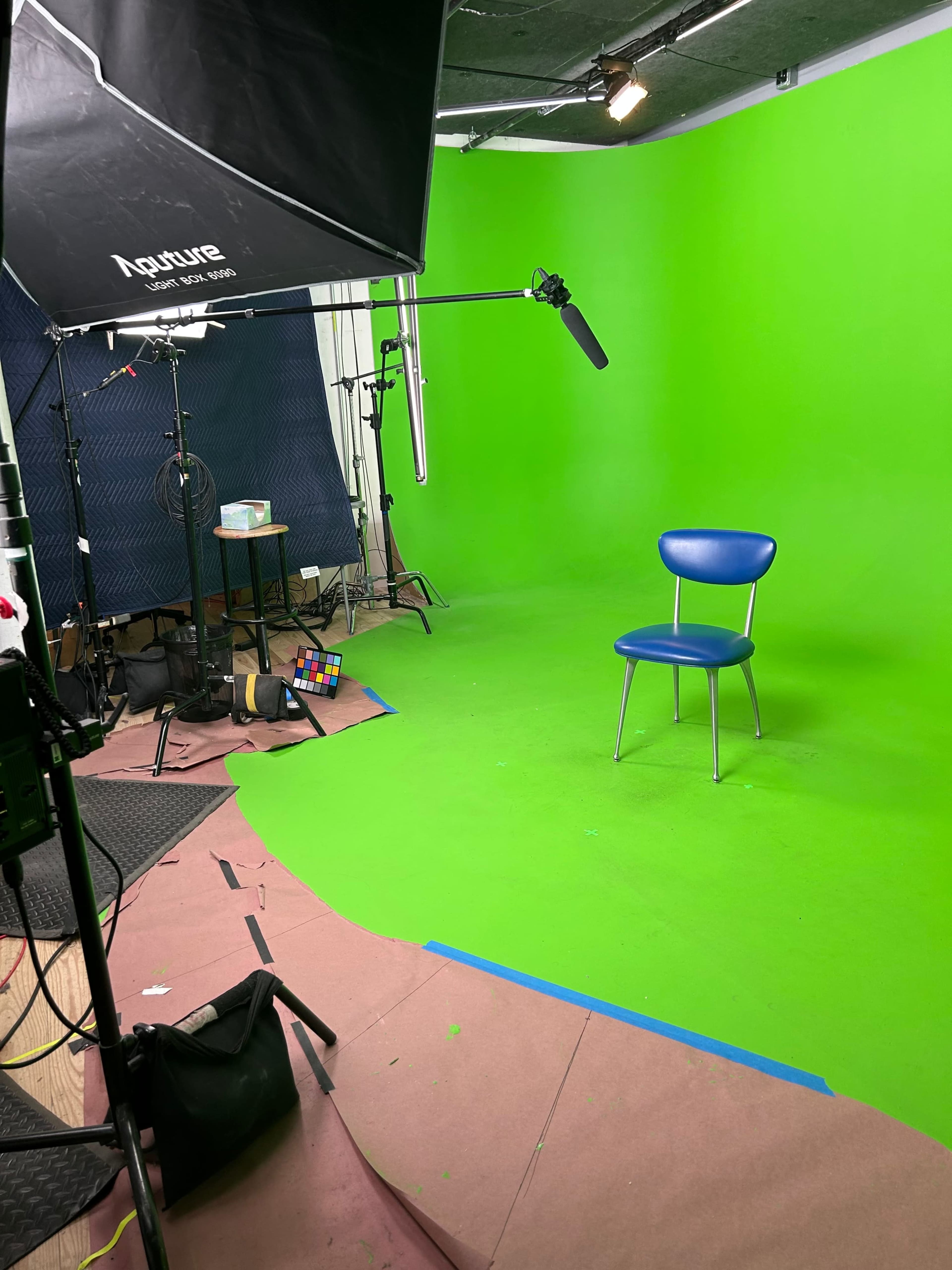 The image shows a green screen studio setup featuring a blue chair in front of a bright green backdrop, with various lighting and camera equipment arranged around the space.