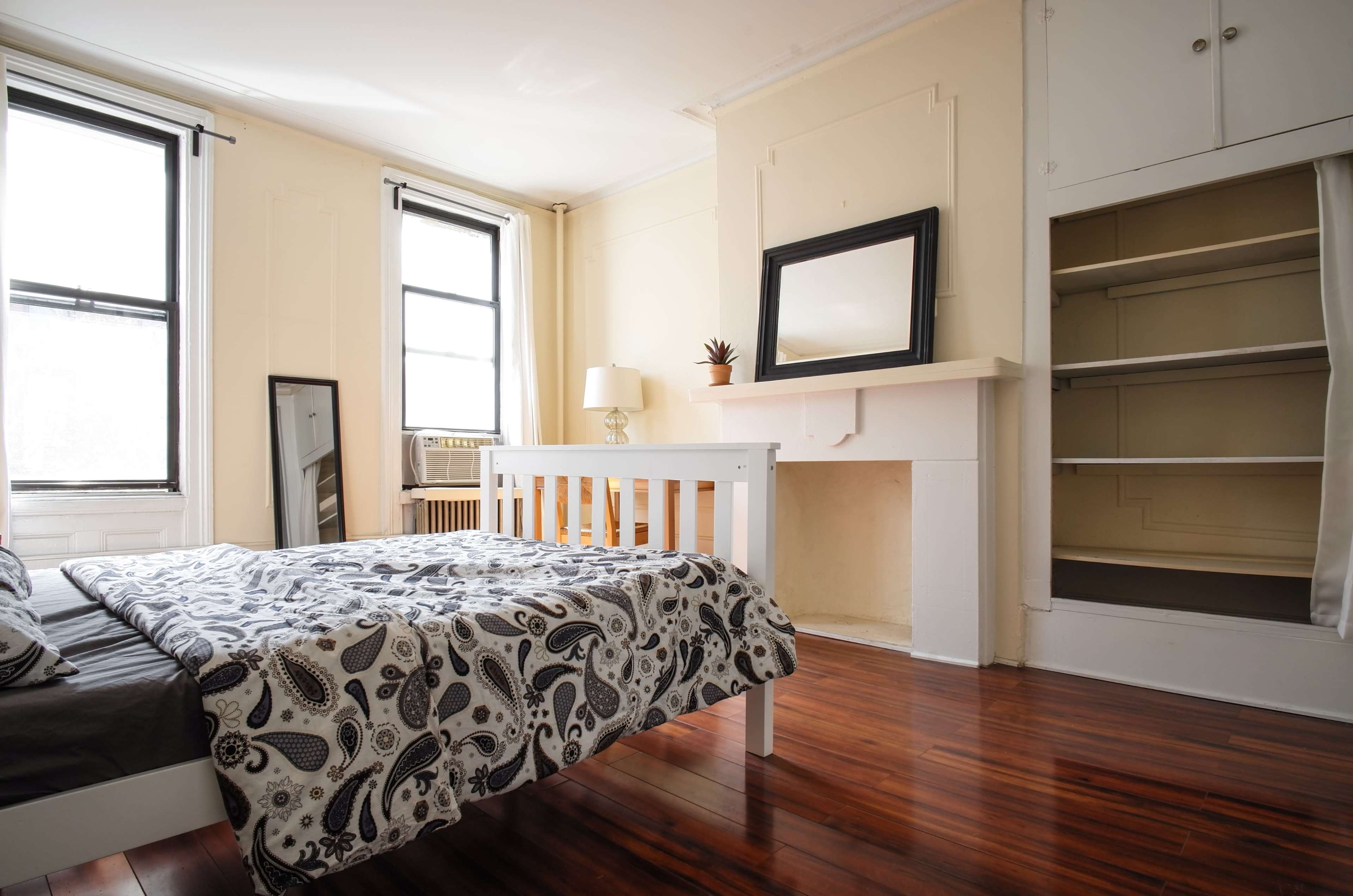 A bedroom with a white bed, hardwood floor, a large mirror, a lamp on a nightstand, and a built-in shelf near a fireplace.