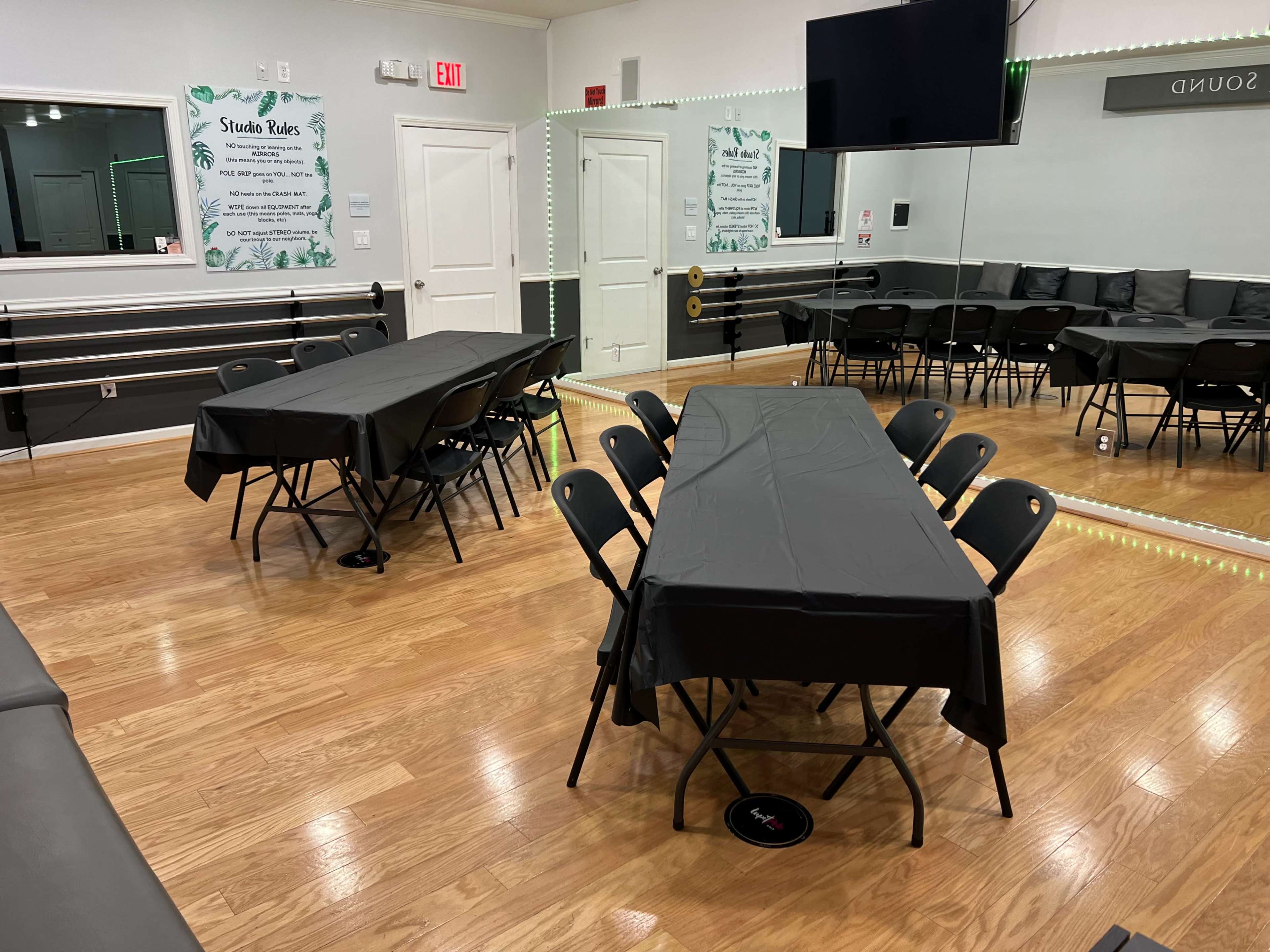 The scene shows a dance studio with two long tables covered in black tablecloths, surrounded by black folding chairs, and mirrors on the walls.