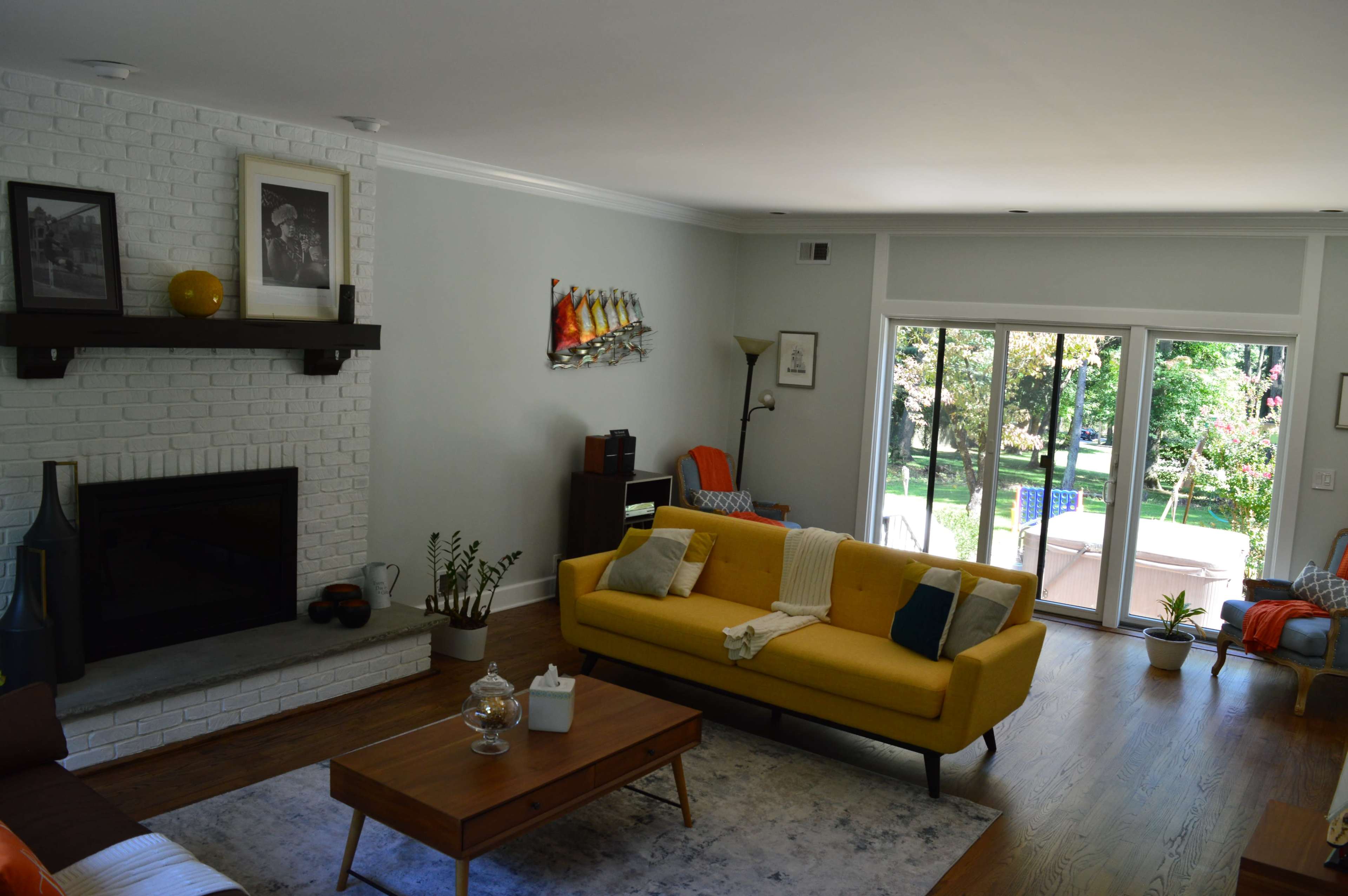 The living room features a yellow sofa, a coffee table, and a fireplace, with large windows that open to a view of a garden.