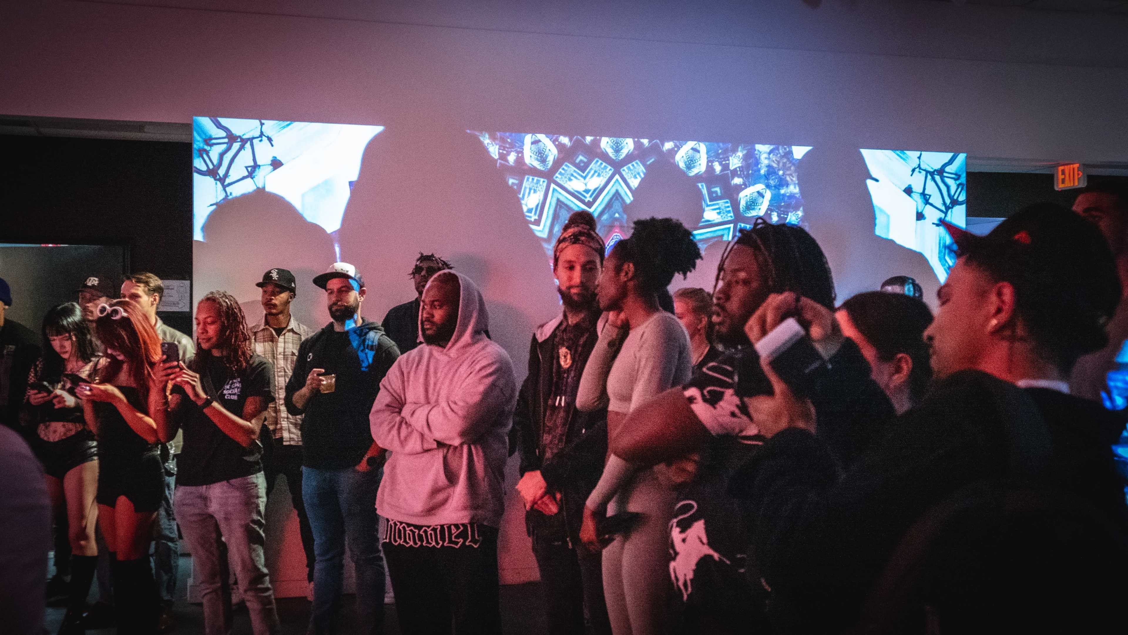A diverse group of people stands in a gallery space with projected visuals on the walls.