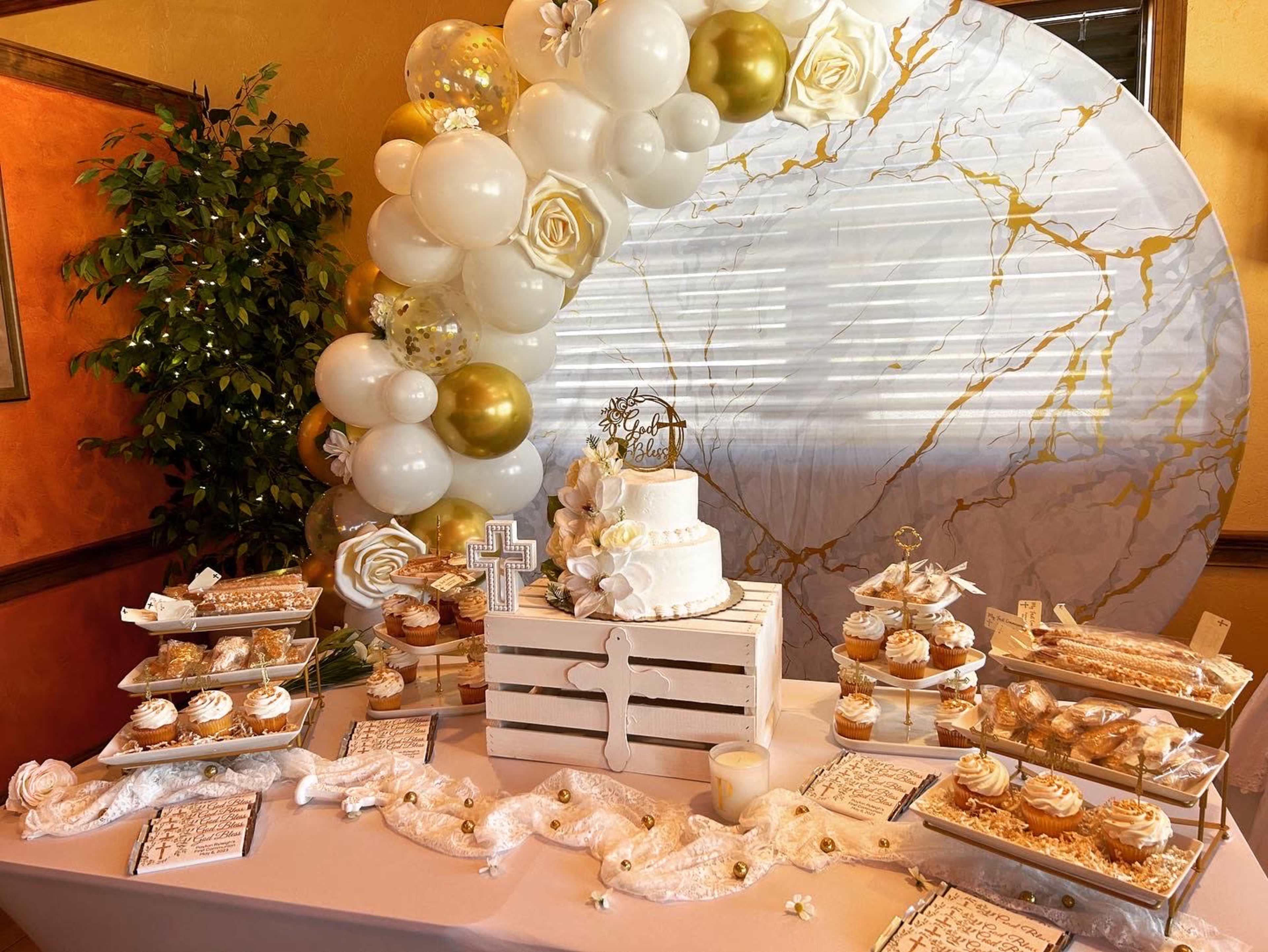 A dessert table displays a tiered cake surrounded by assorted pastries, decorated with balloons and floral elements against a backdrop of a golden marble design.