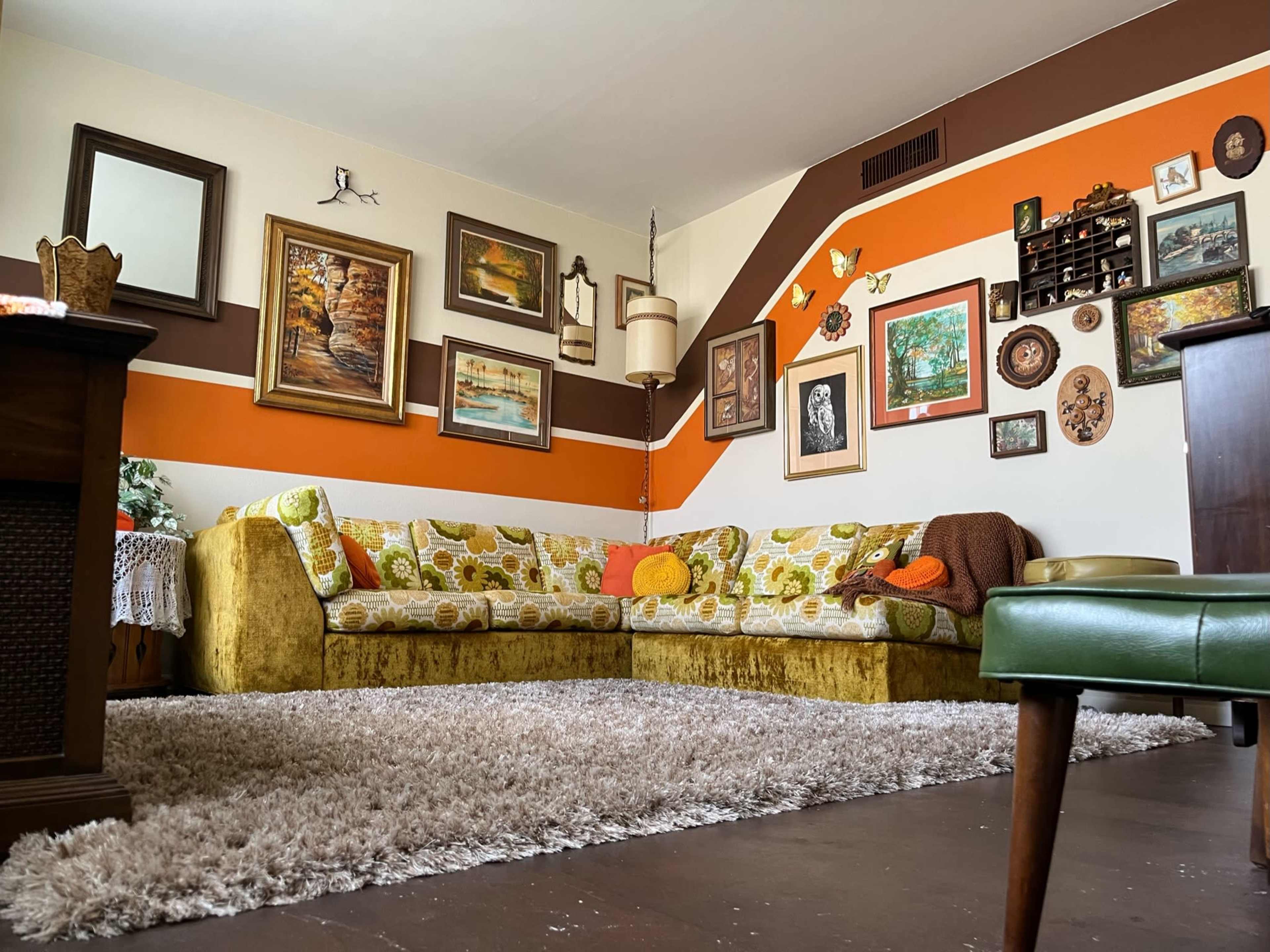 The image shows a living room with a large, patterned sectional sofa, a shaggy rug, and various framed artworks on the walls.