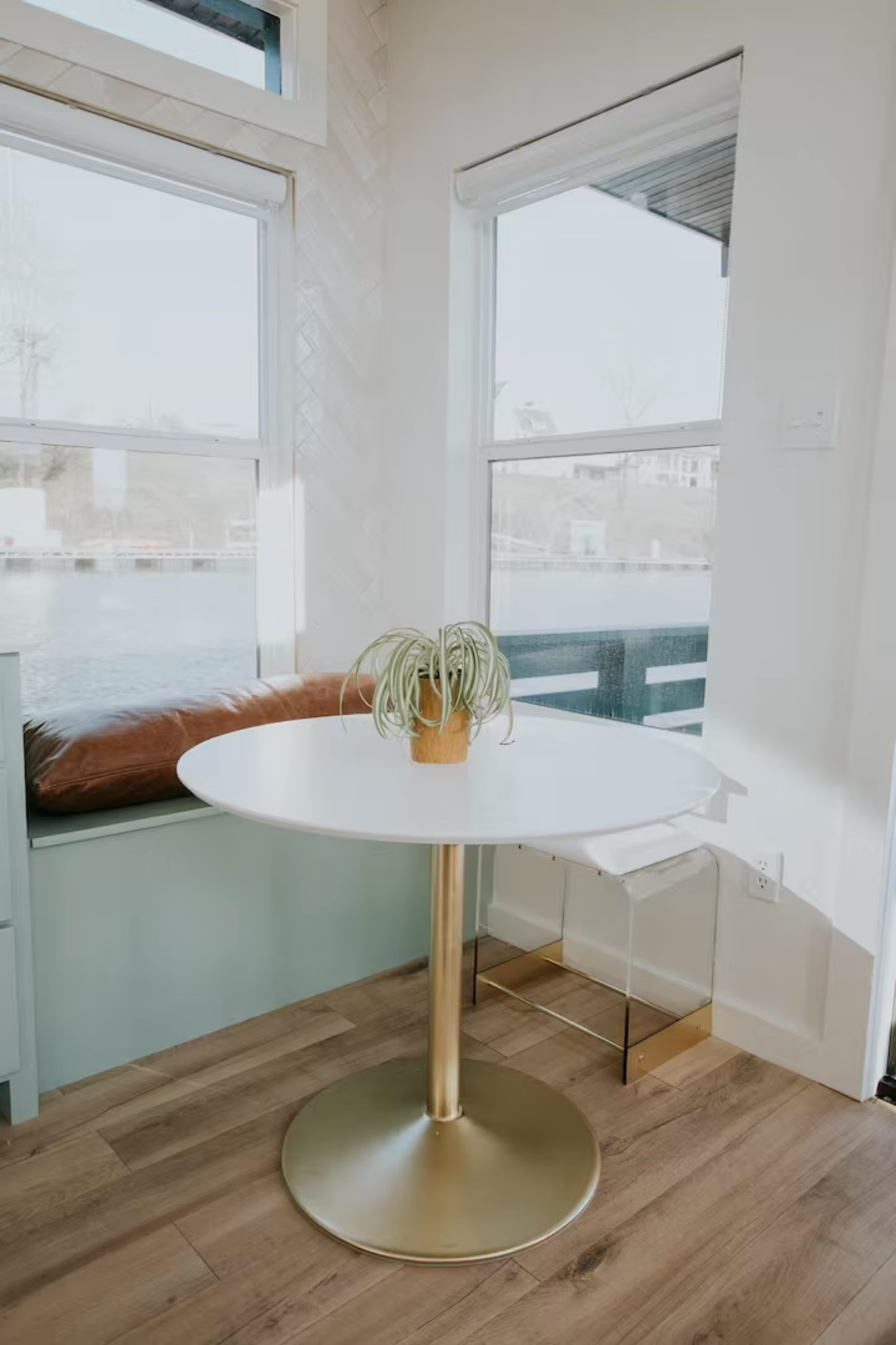 A round white table with a gold base is placed near a window, accompanied by a potted plant, and a cushioned bench is situated nearby.
