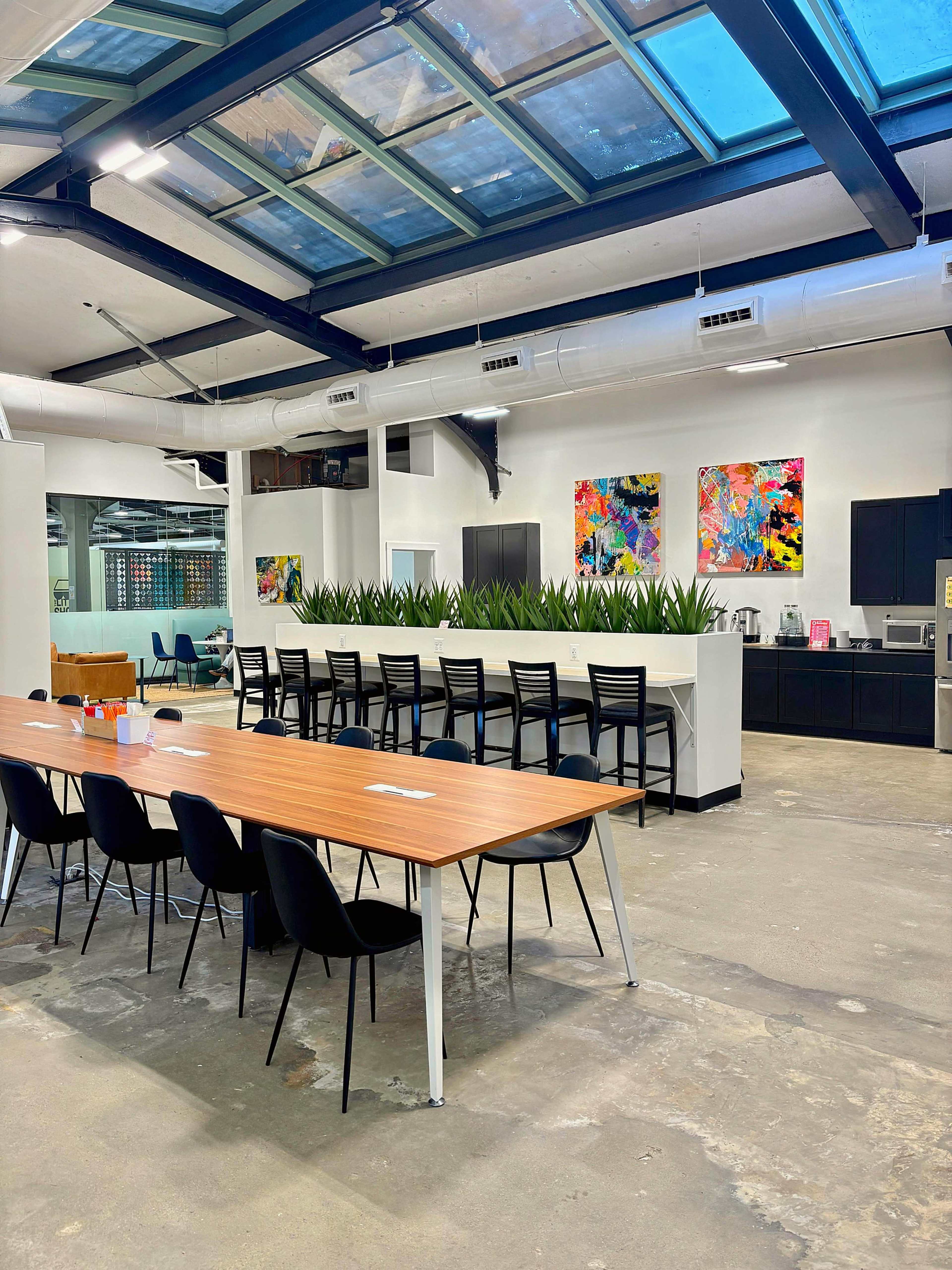 The image shows a modern office kitchen and dining area with a long wooden table, black chairs, and vibrant artwork on the walls, illuminated by natural light from the skylights above.