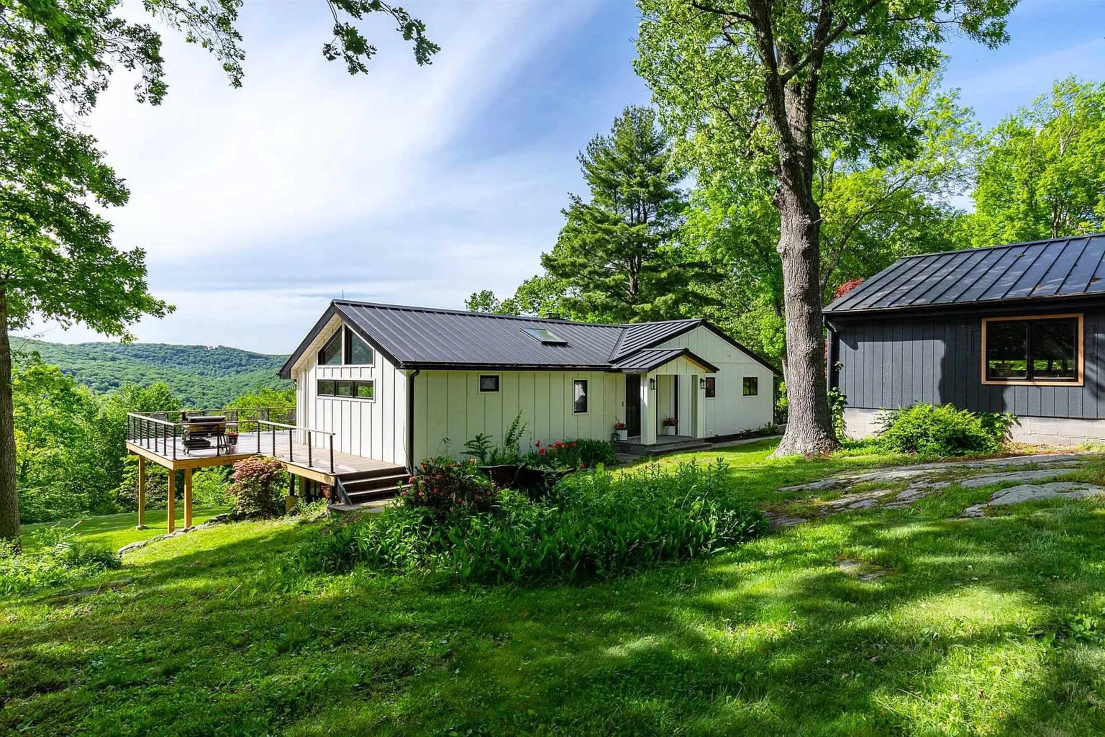 A white and black house sits on a grassy hill surrounded by trees, with a deck and a scenic view of the distant landscape.