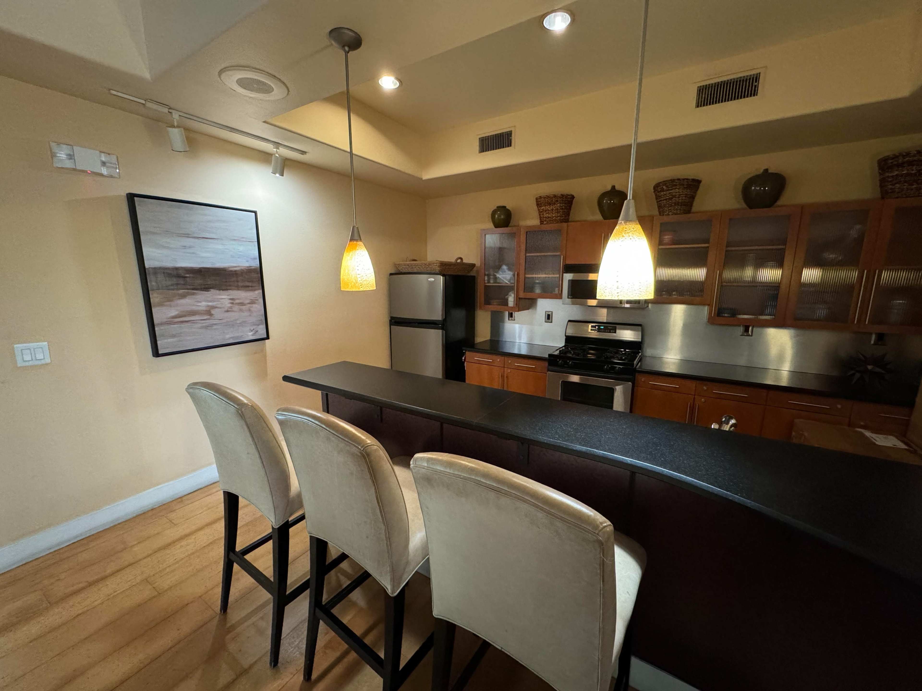 A modern kitchen features a bar with three stools, overhead lighting, and stainless steel appliances.