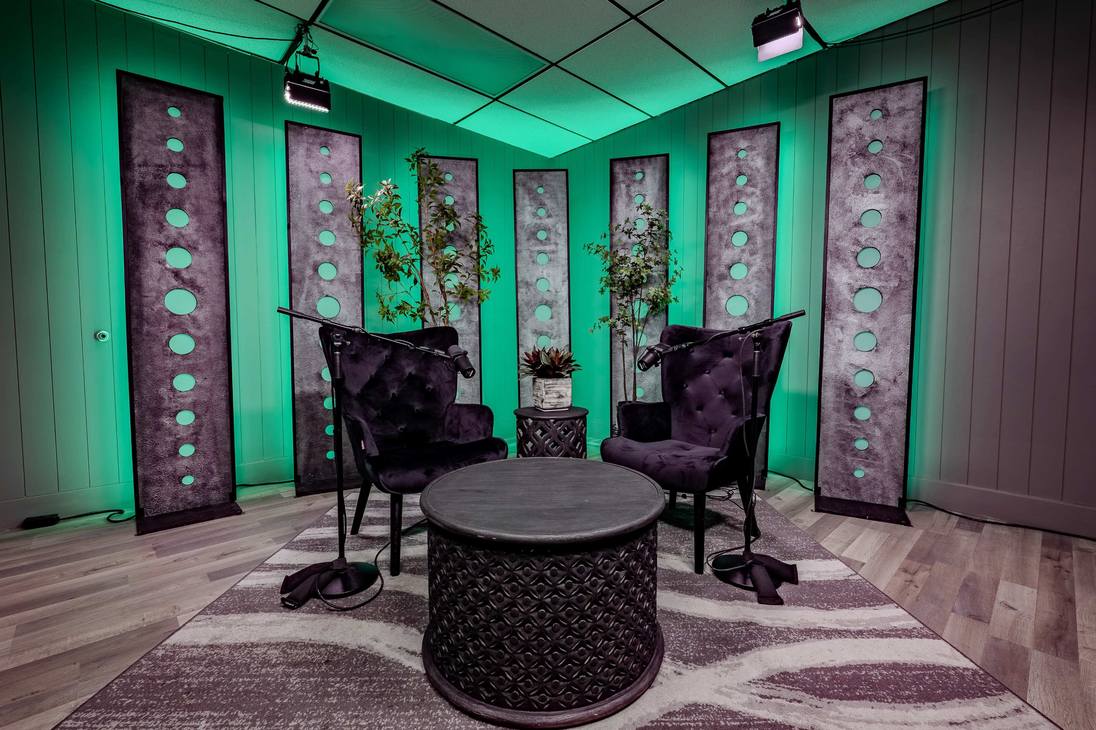The image shows a modern recording studio setup with two plush chairs facing a round table, surrounded by tall panel backdrops with circular cutouts and green lighting.