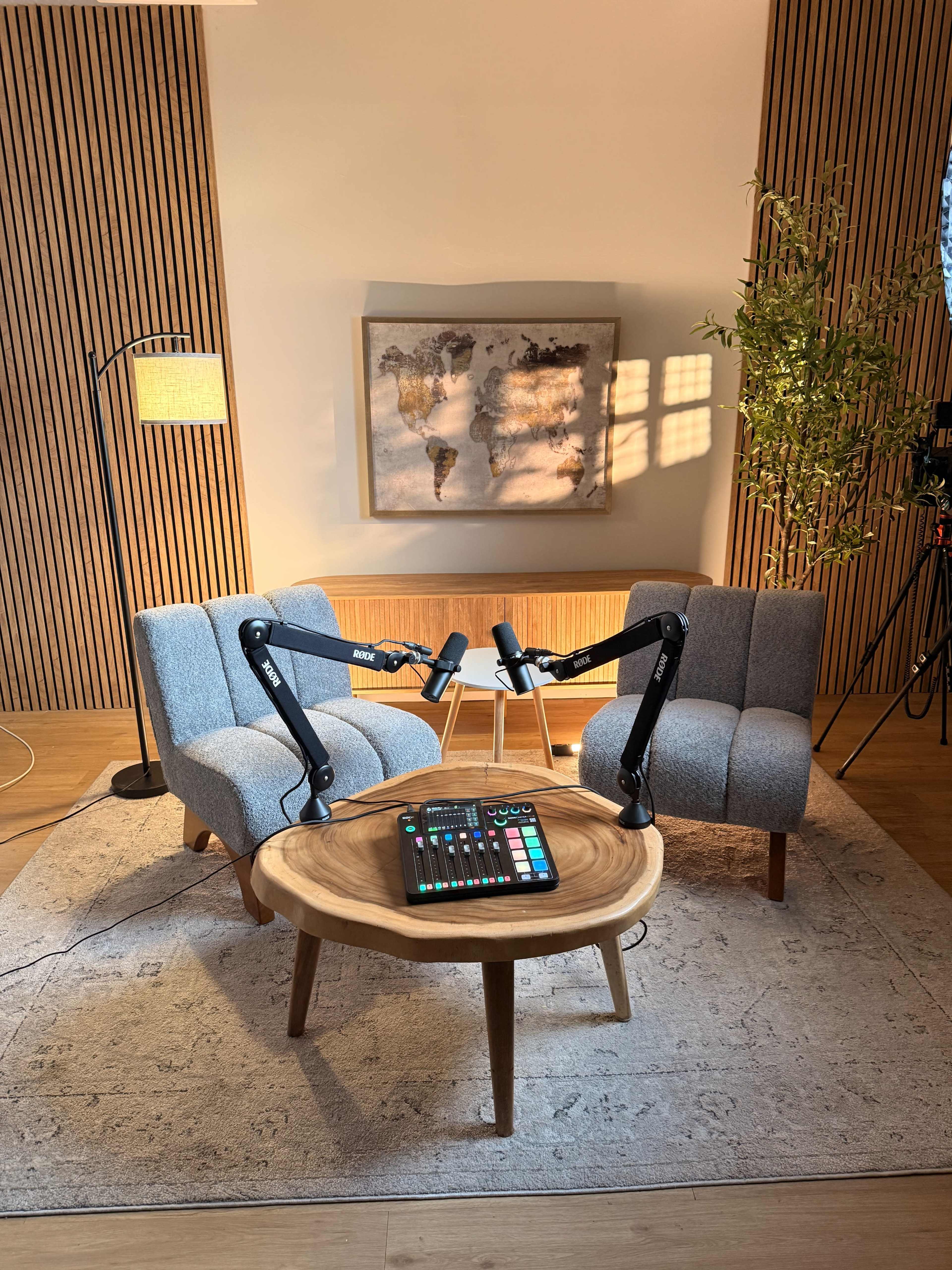 The image depicts a podcast recording setup featuring two armchairs, a round wooden table with audio equipment, and a painting on the wall in a well-lit room.