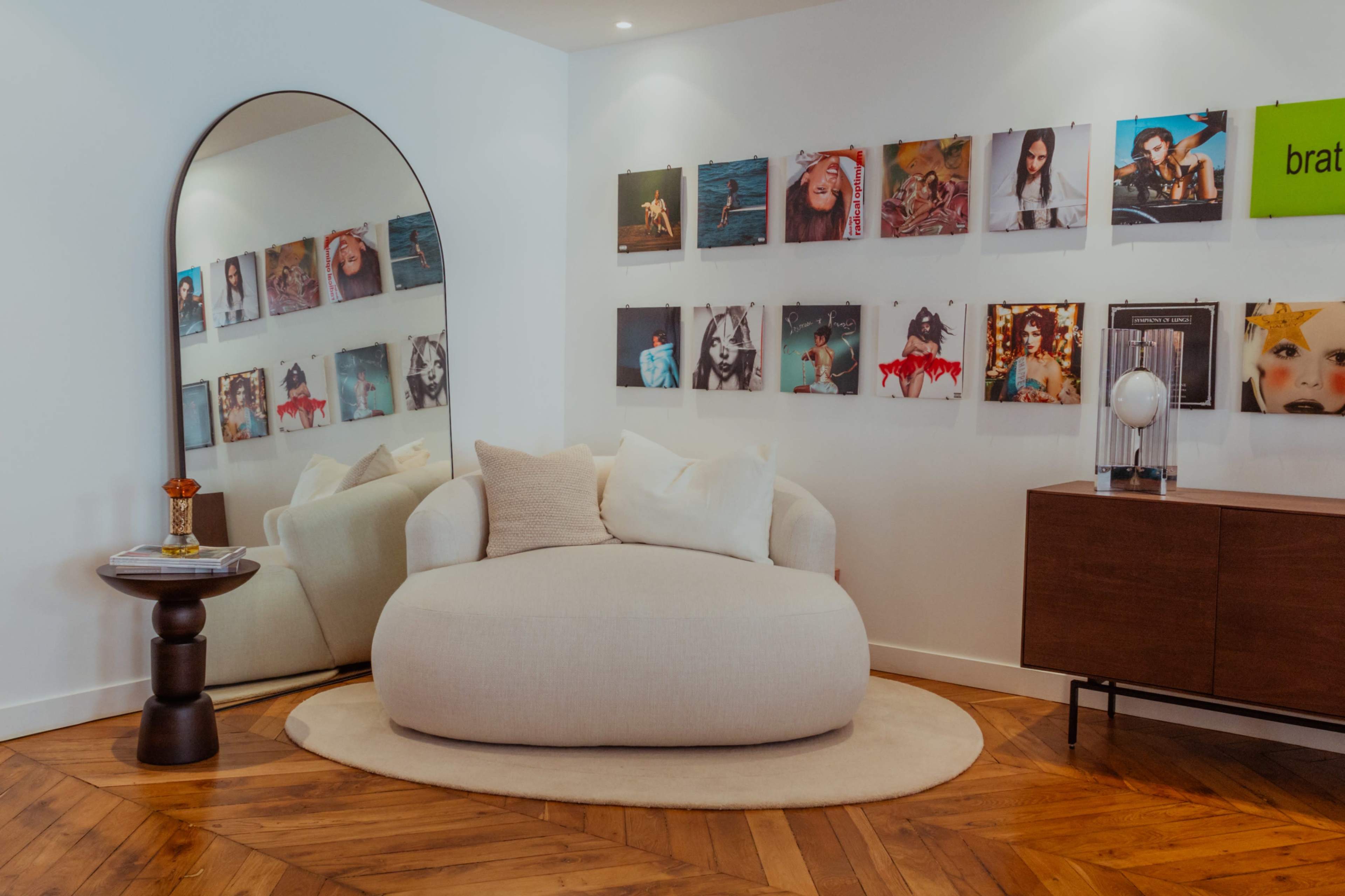 The room features a curved beige sofa, a round table, a mirror, and a gallery wall adorned with colorful photo prints.
