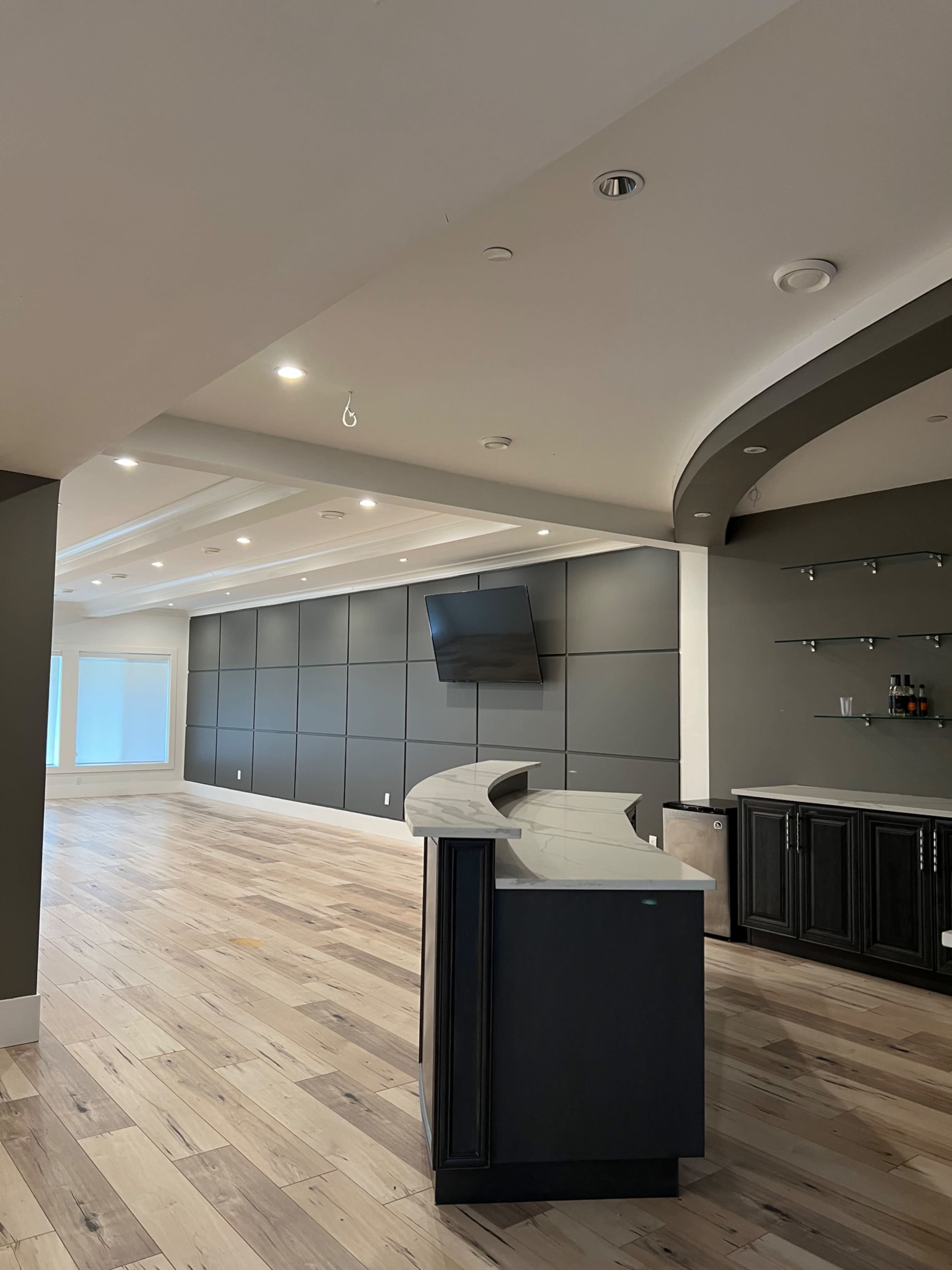 A spacious room featuring a curved bar area with a dark countertop, a wall-mounted television, and a section with shelving, all complemented by wooden flooring.
