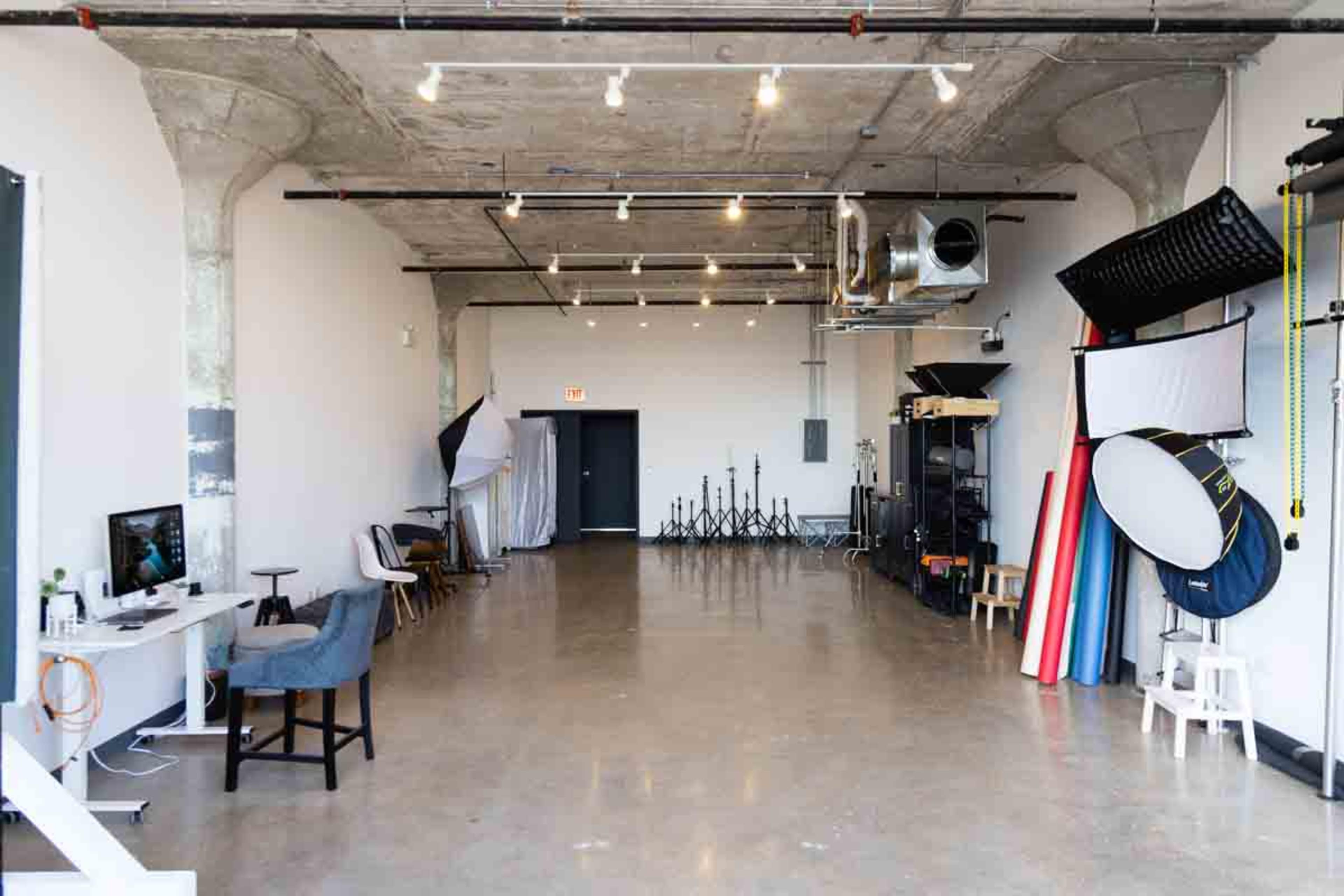 The image shows a spacious photography studio with various lighting equipment, backdrops, and a computer workstation set against a minimalist concrete wall.