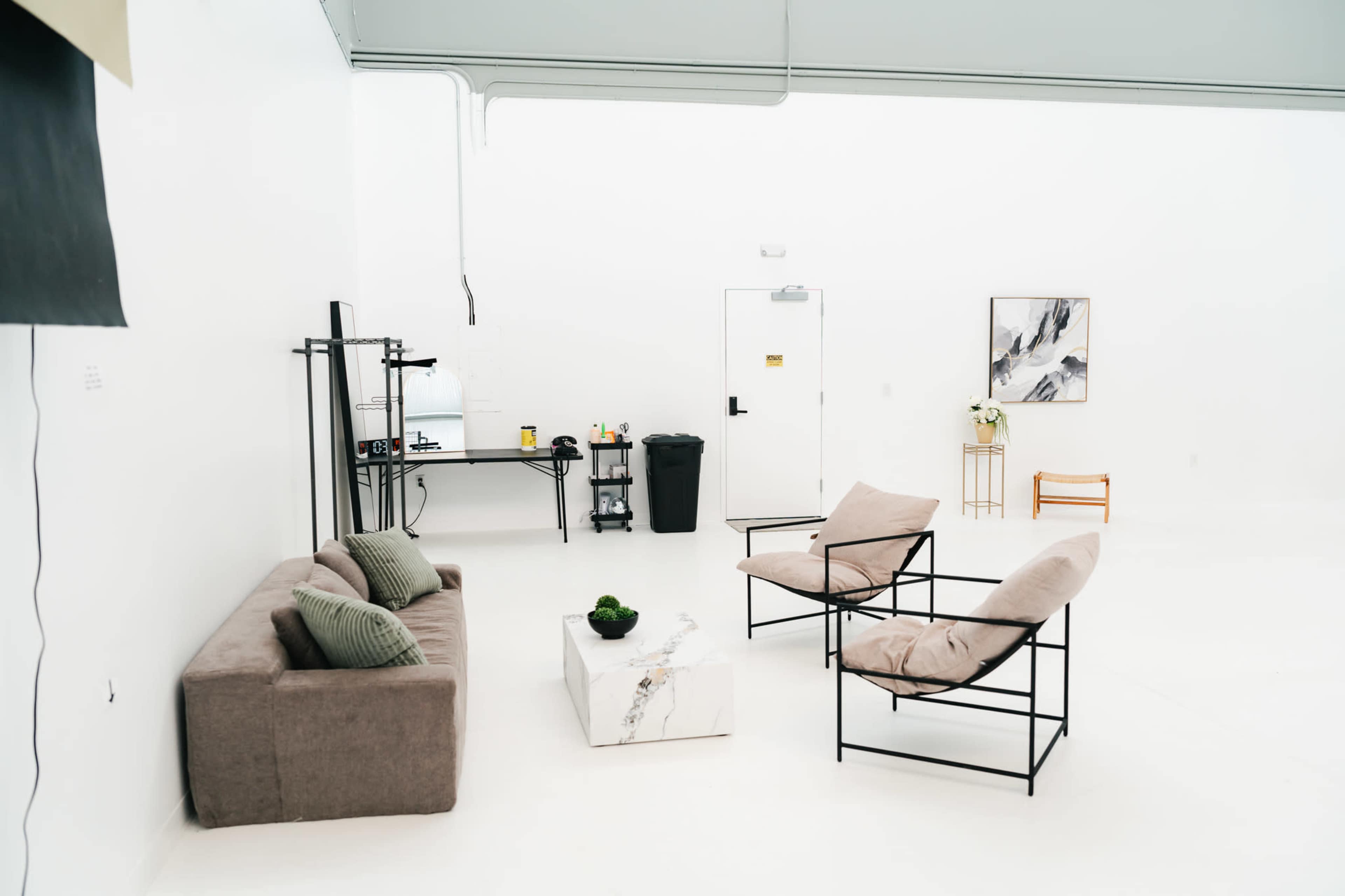 The image shows a spacious, minimalist room with a sofa, two armchairs, a coffee table, a small kitchen setup, and a few decorative elements.